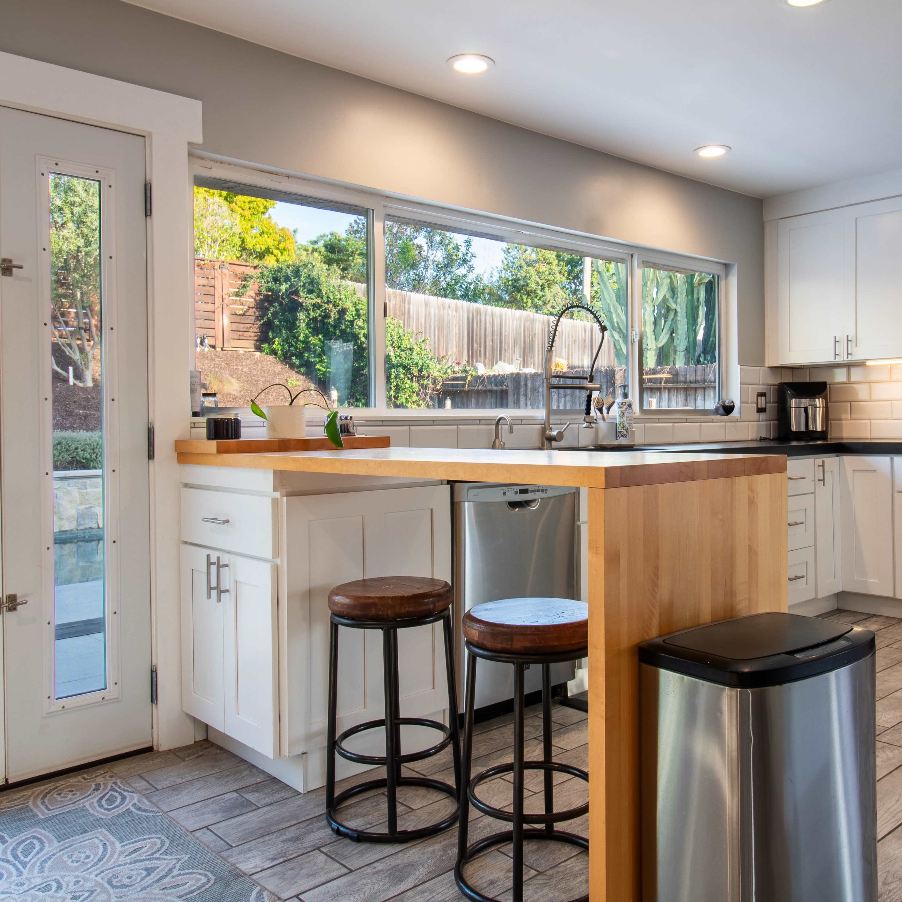 A modern kitchen features a central island with stools, stainless steel appliances, and large windows overlooking a backyard pool area.
