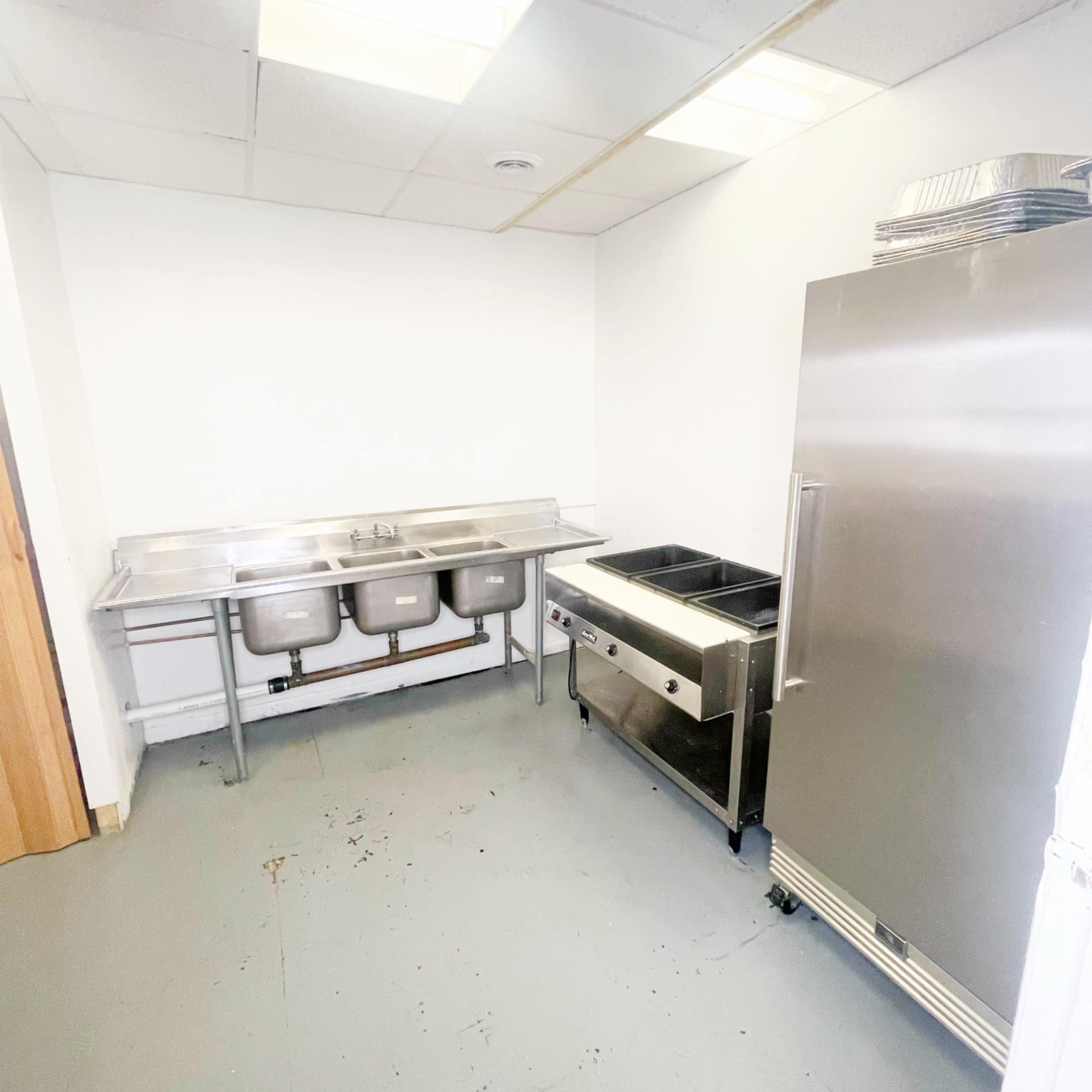 A sparse commercial kitchen with a stainless steel sink, a refrigerated unit, and a countertop covered with a serving station.