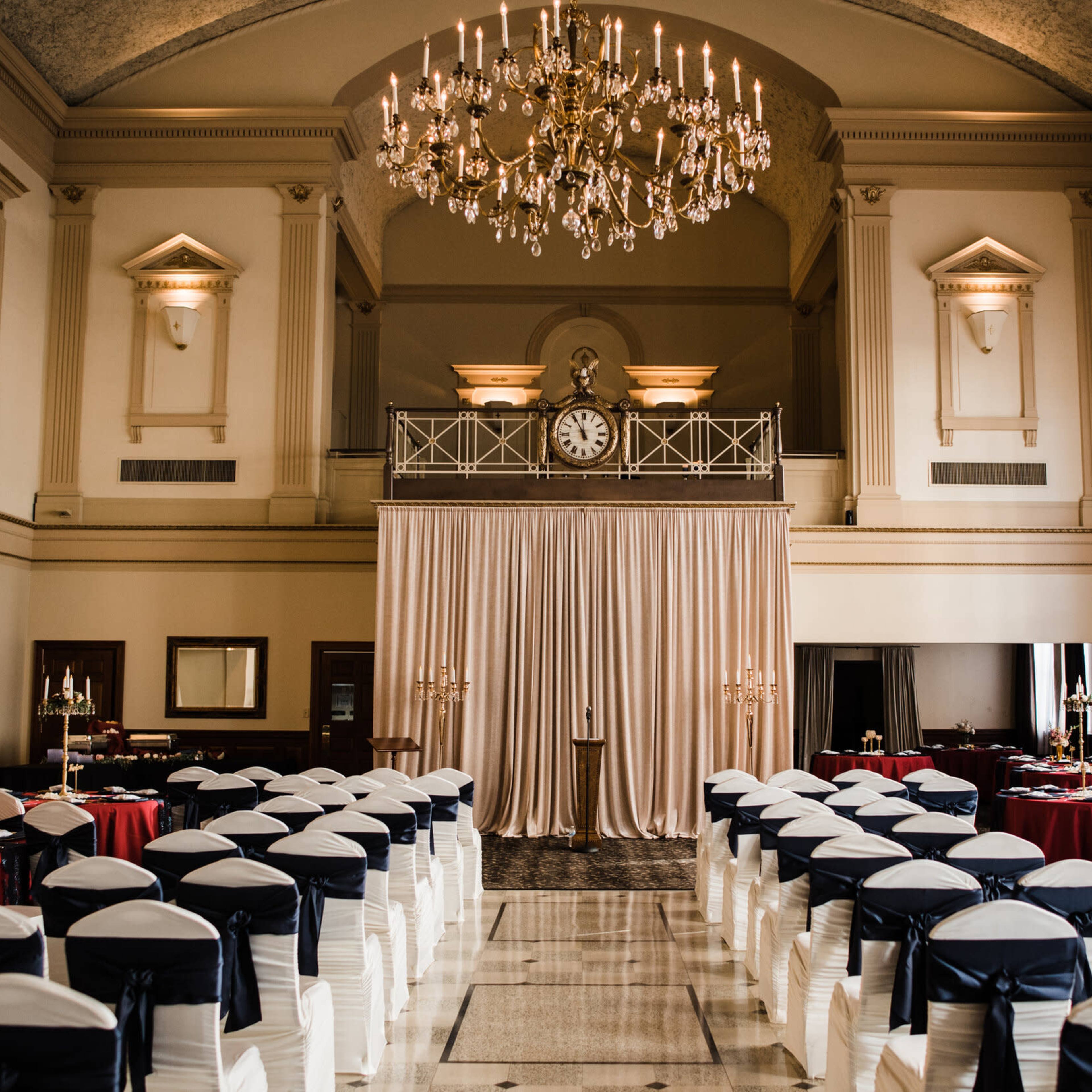 A spacious banquet hall set for an event, featuring rows of chairs draped in white with blue sashes, elegant tables adorned with red tablecloths, and a large chandelier overhead.