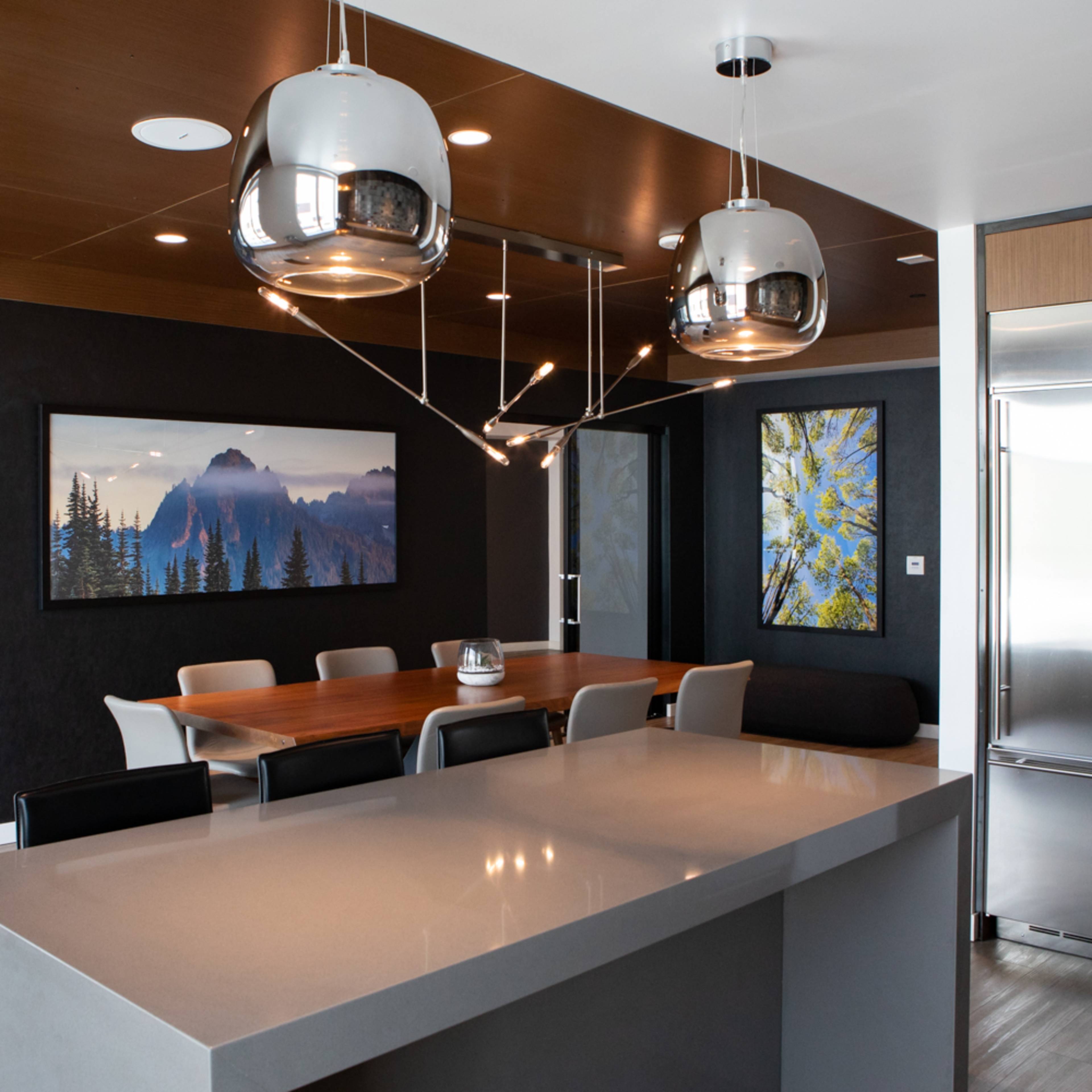 A modern kitchen features a central island with a gray countertop, a dining area with a wooden table surrounded by chairs, and artwork on the walls.