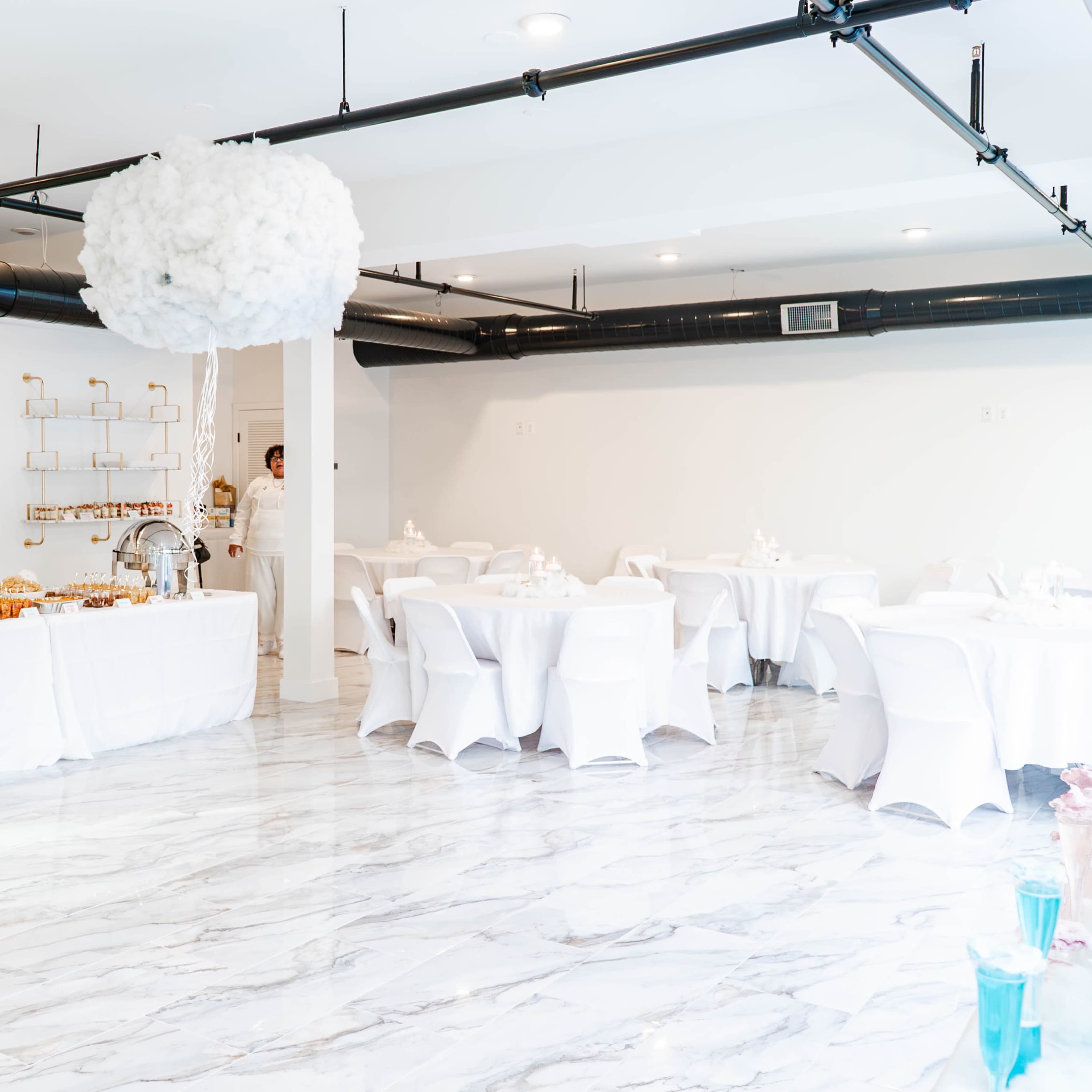 A spacious event room features white tablecloths, round tables set for guests, and a buffet table with various food items against a light-colored backdrop.