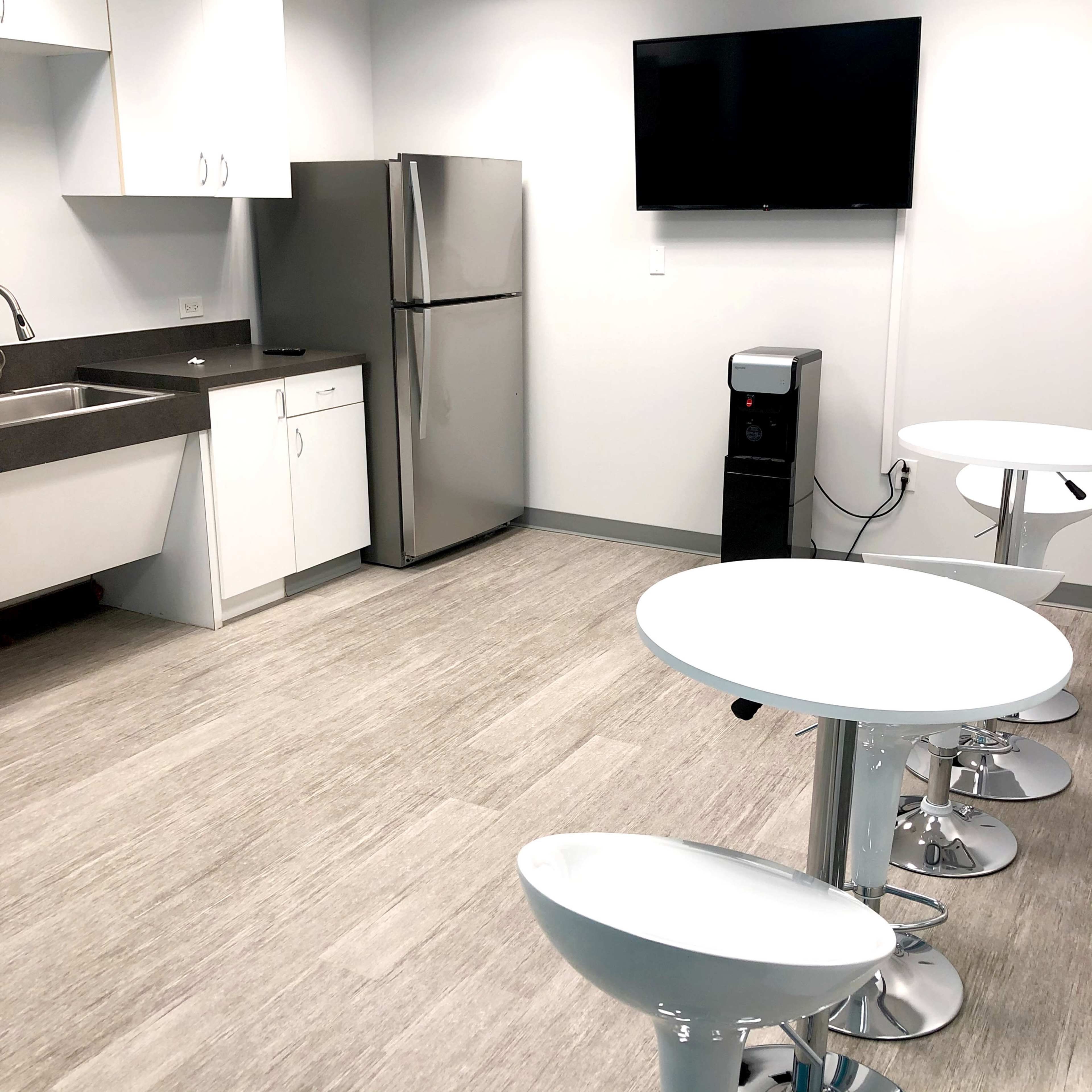 A modern kitchen area with a refrigerator, sink, and a TV mounted on the wall, accompanied by two round tables and four bar stools.