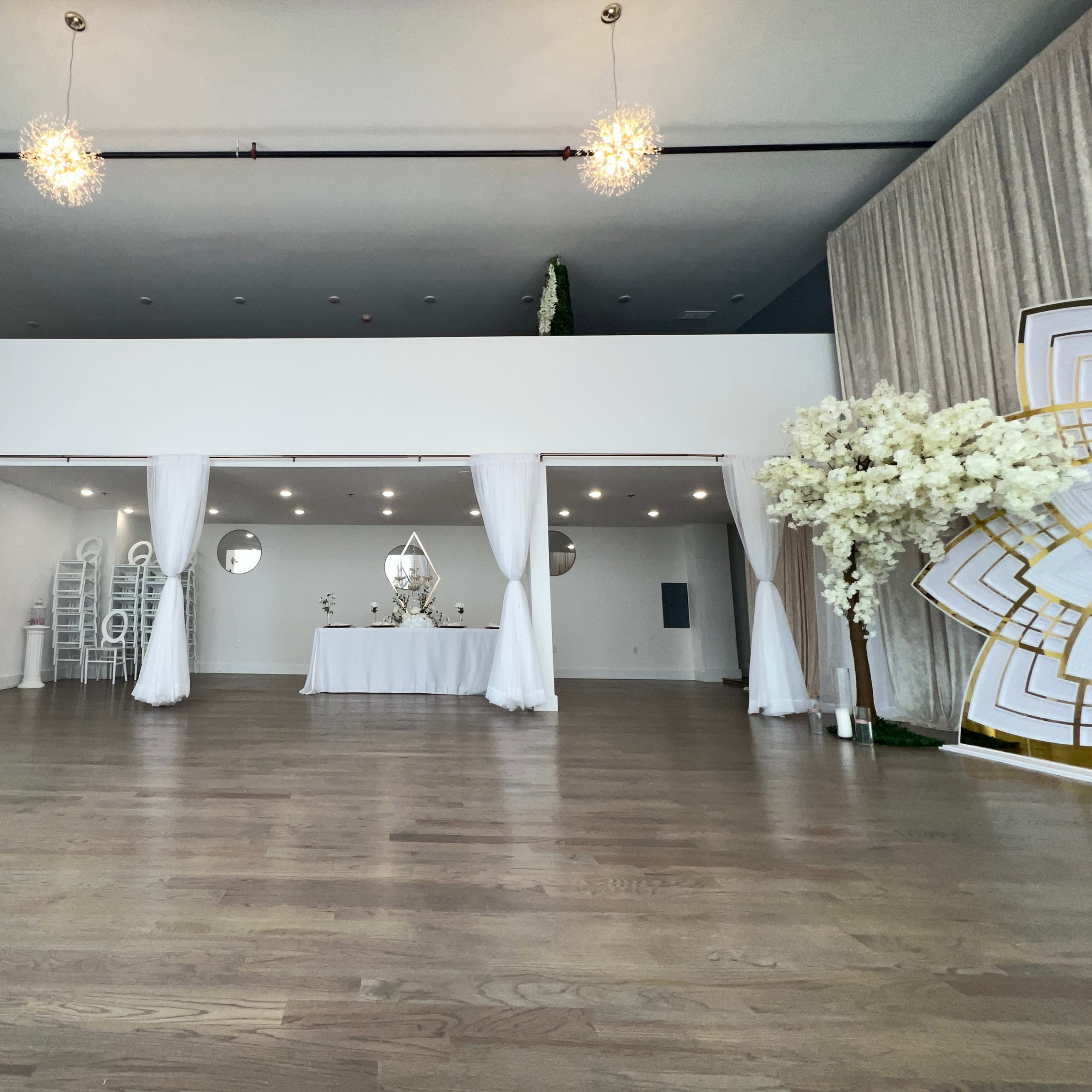 A spacious interior venue with a white draped area, a table set at the center, and elegant decor elements including floral arrangements and lighting fixtures.