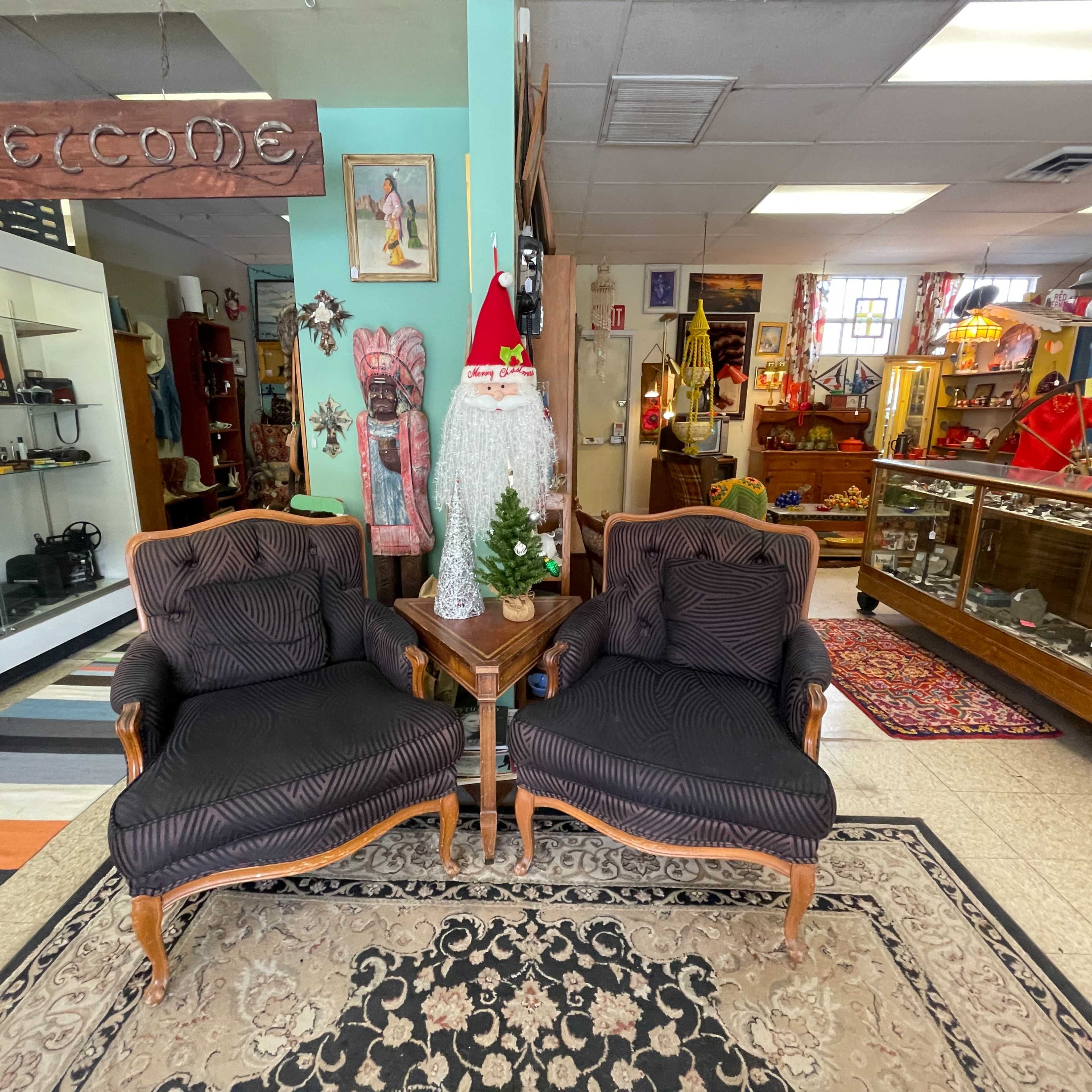 A vintage shop interior with two upholstered chairs, a small table, and various decorations including a Christmas-themed figure and shelves filled with items.