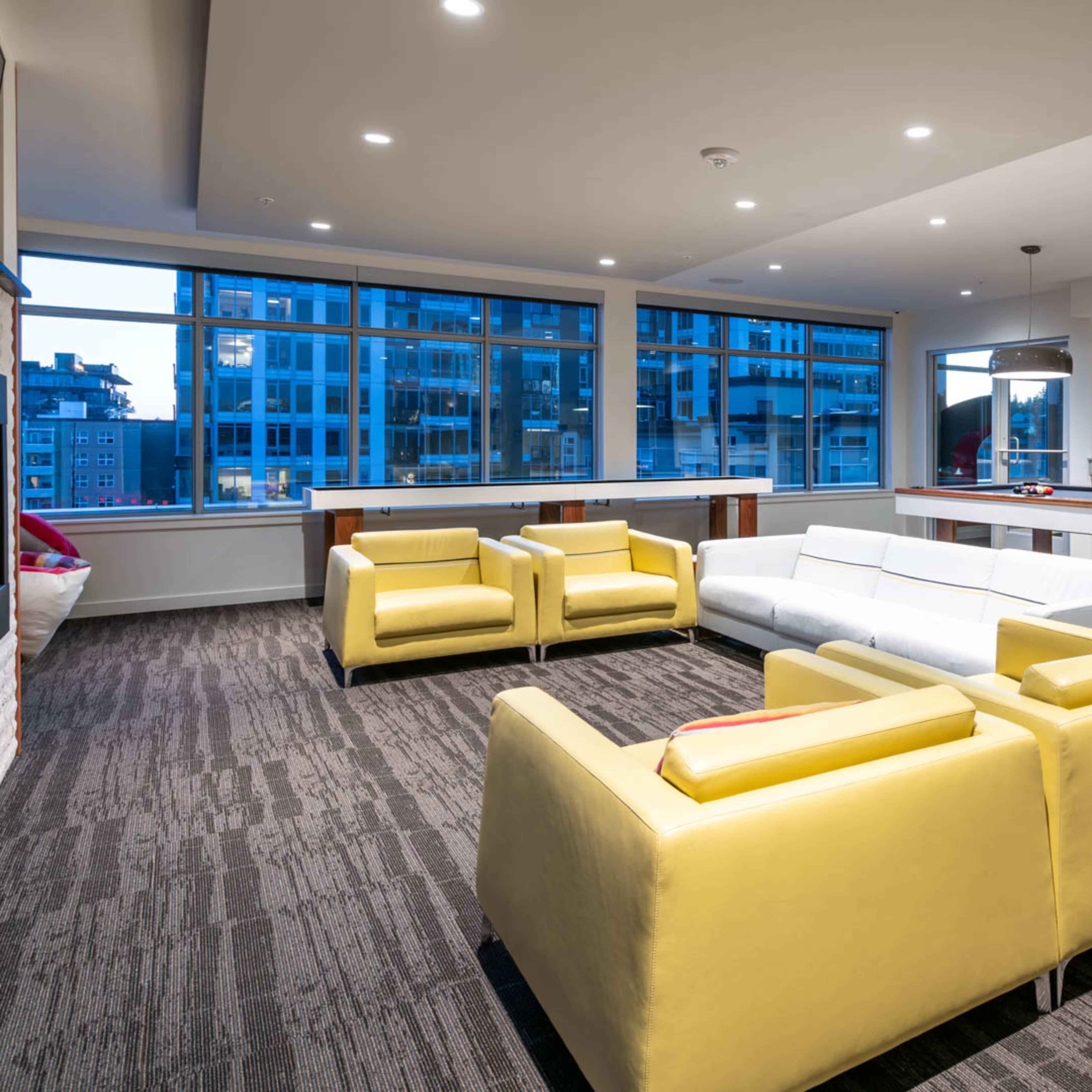 A modern lounge area with yellow and white seating, large windows revealing a cityscape, and a kitchen area in the background.