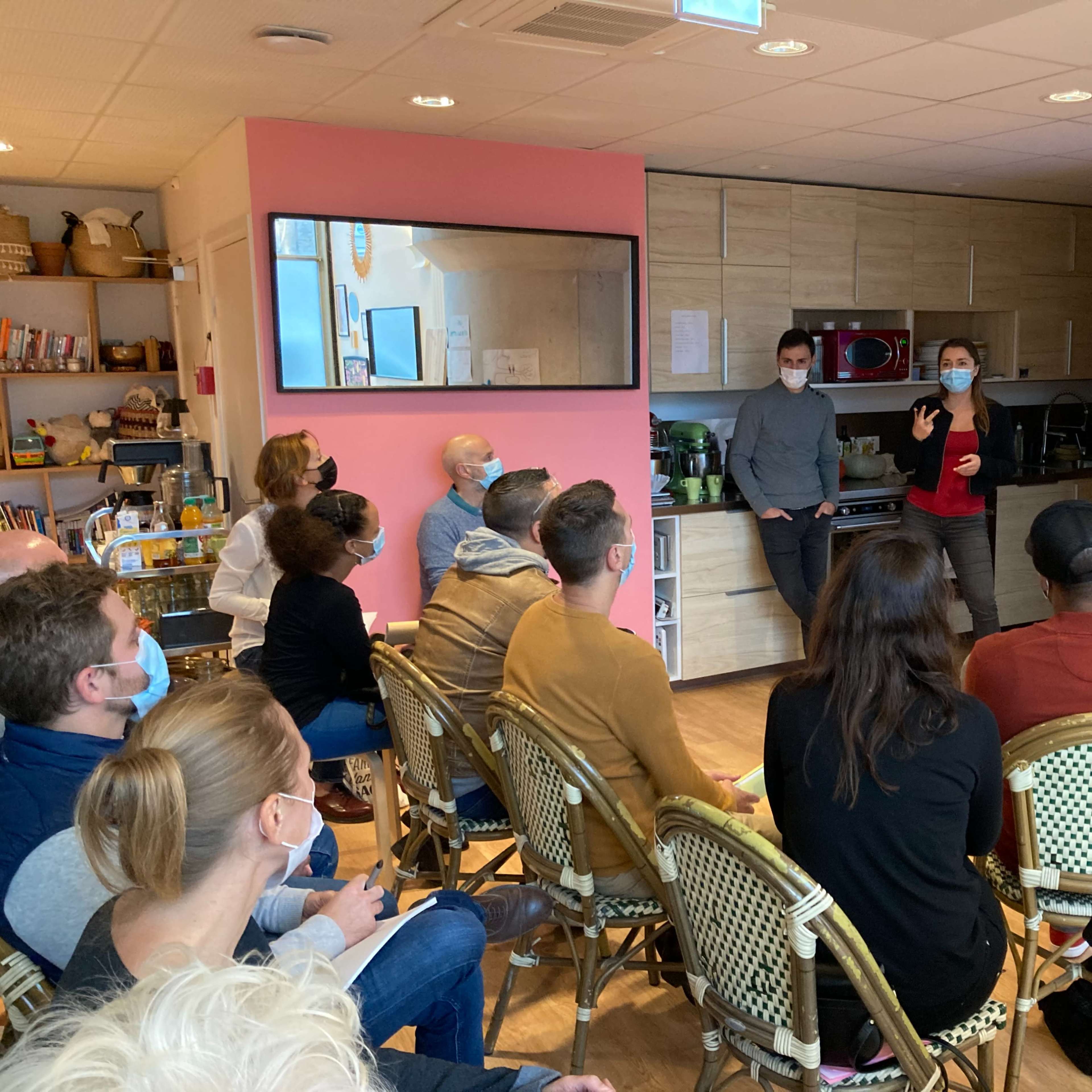 A group of people is seated in a kitchen area, listening to a presentation projected on a screen.