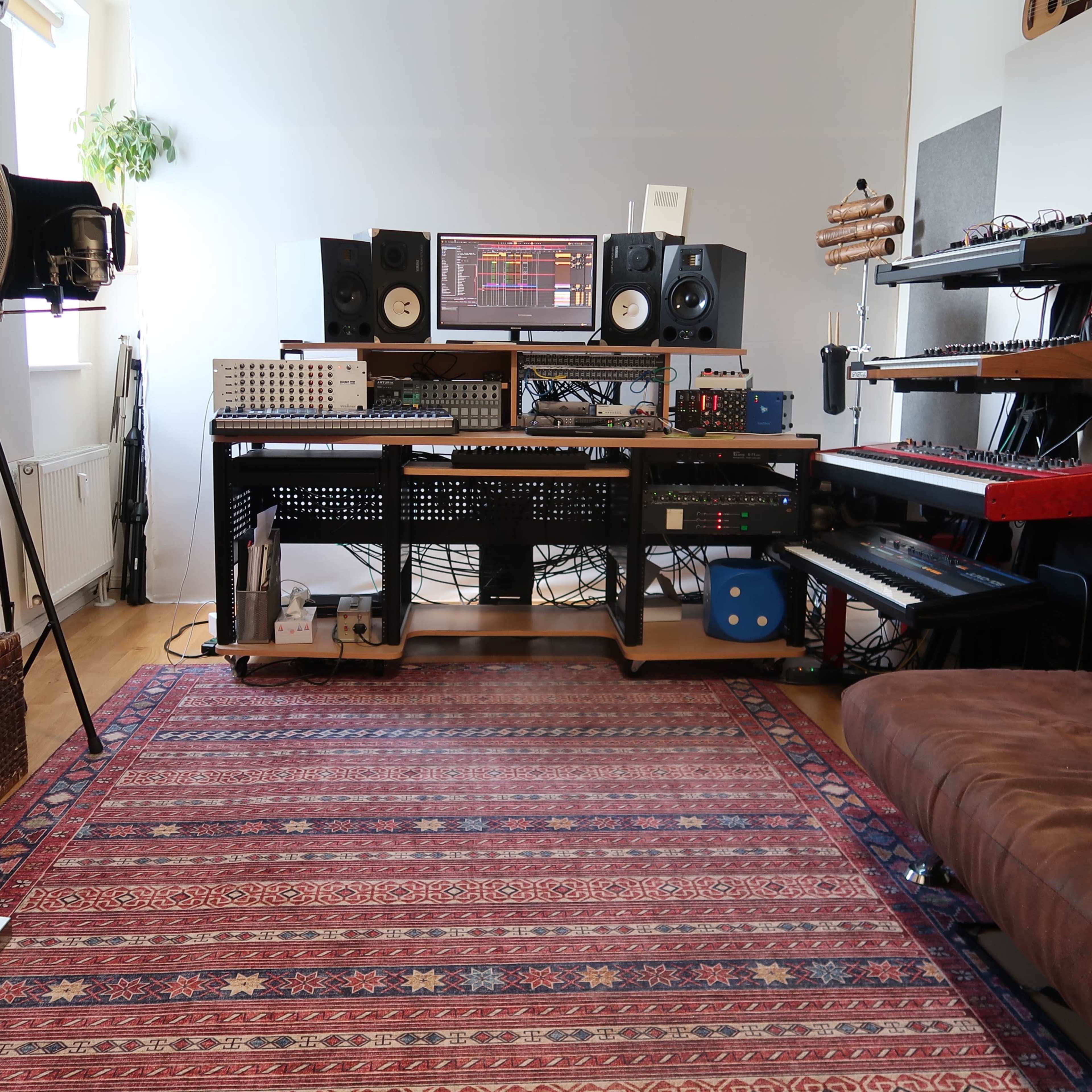 A music studio setup with a desk featuring equipment, speakers, and a computer monitor, alongside a couch and a patterned rug.