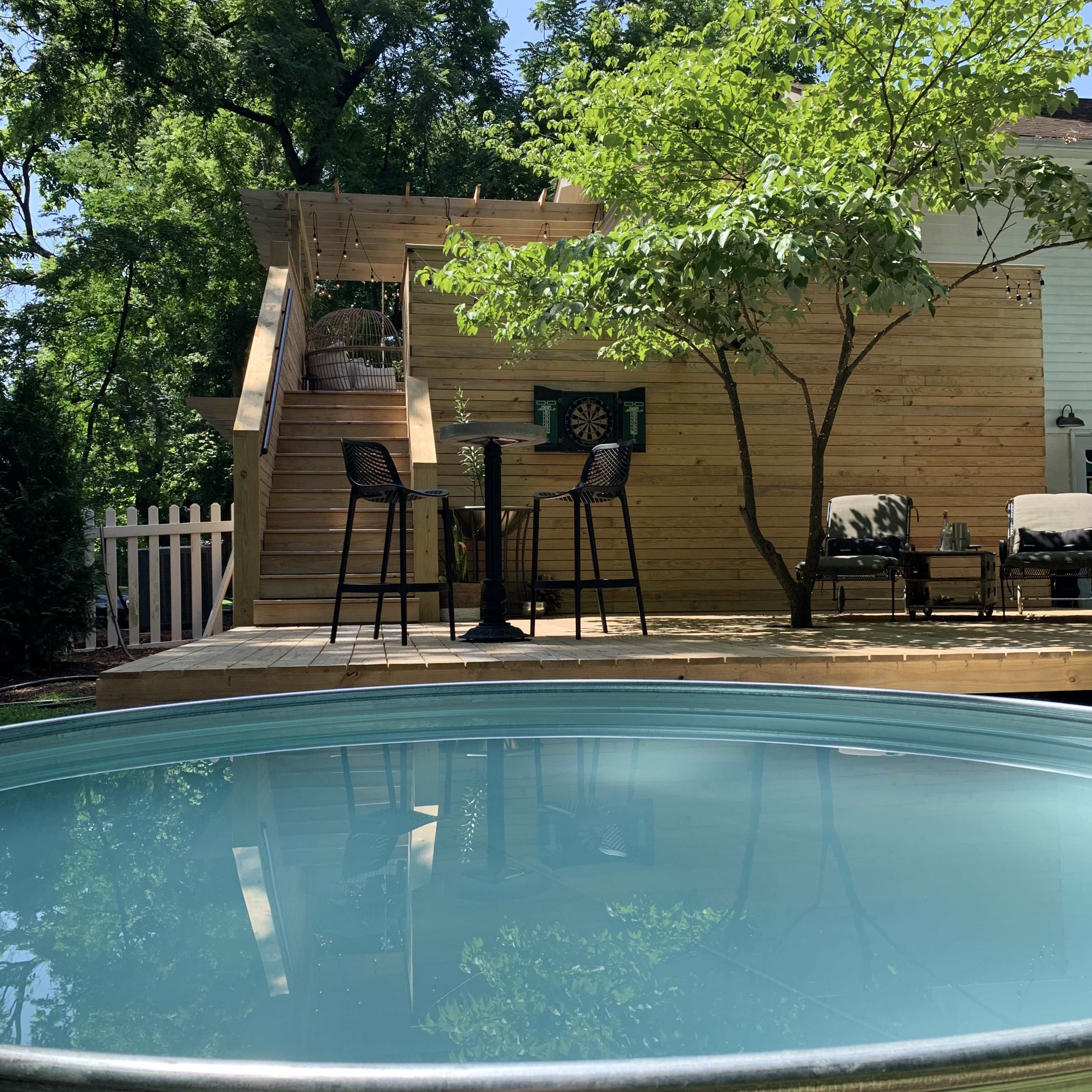 A circular swimming pool reflects a sunlit wooden deck with a tree and a staircase leading to an elevated area furnished with chairs and a dartboard.