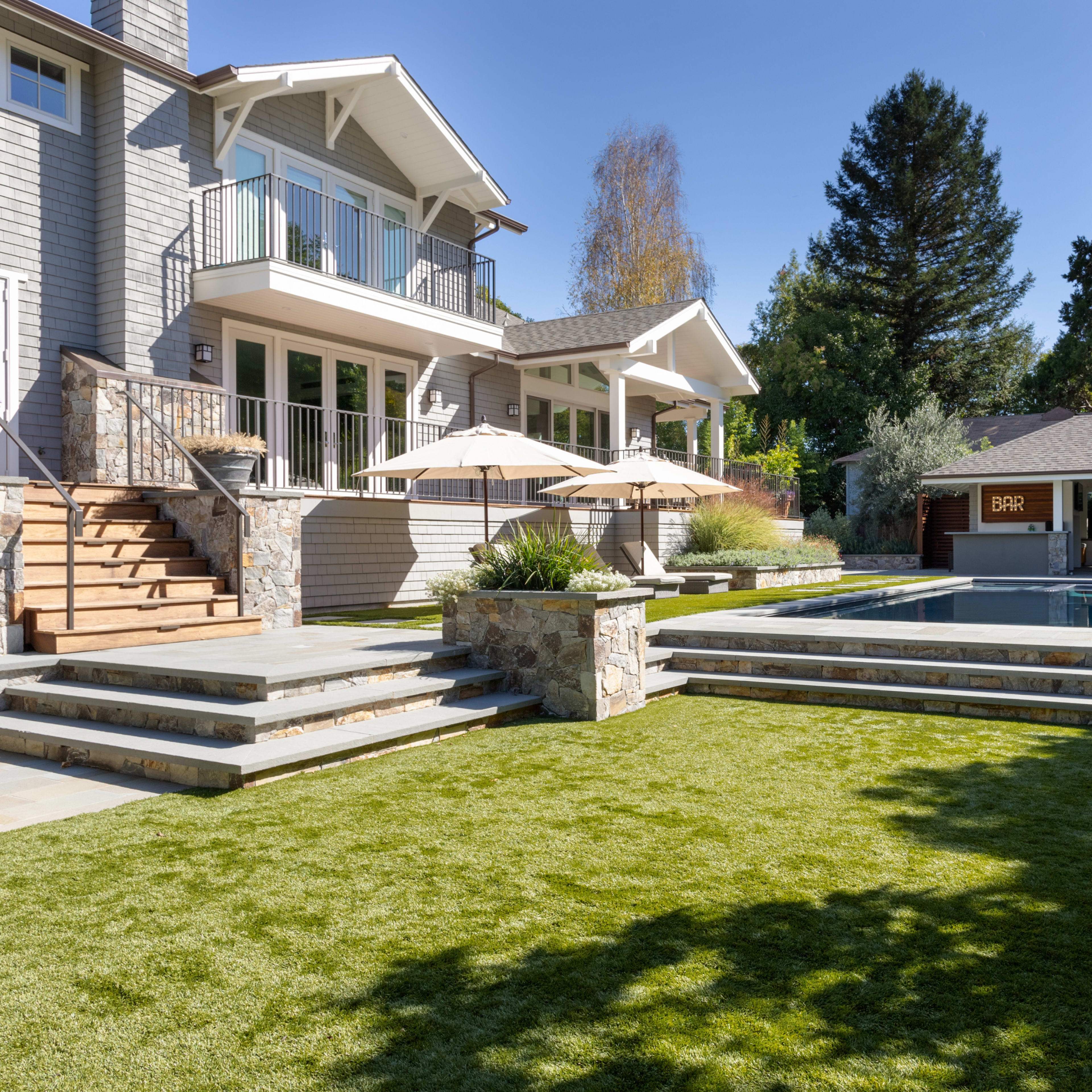 A modern home with a landscaped yard, featuring a swimming pool, stone steps, and patio umbrellas.