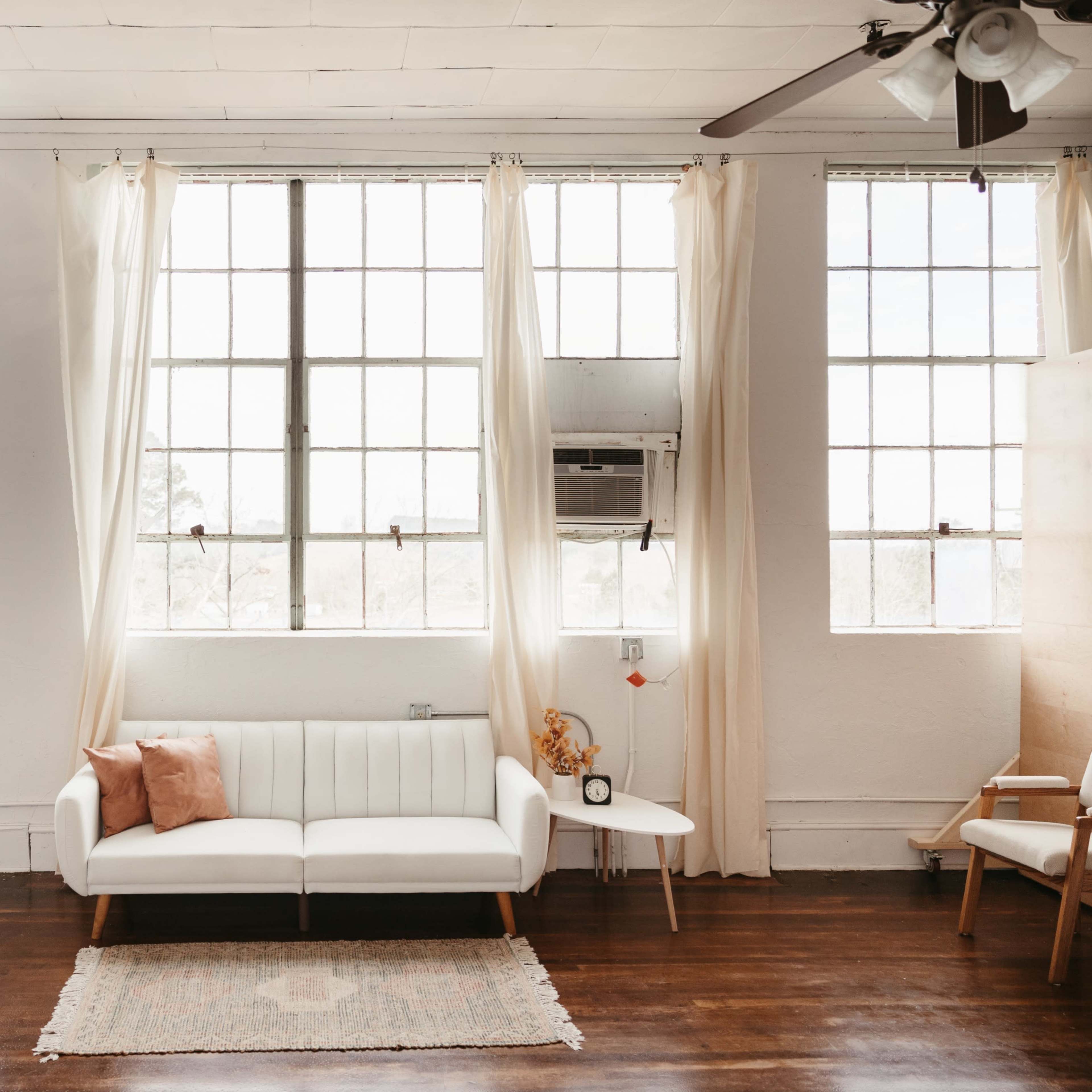 A bright room features large windows, a white sofa, a small side table, and hardwood floors.