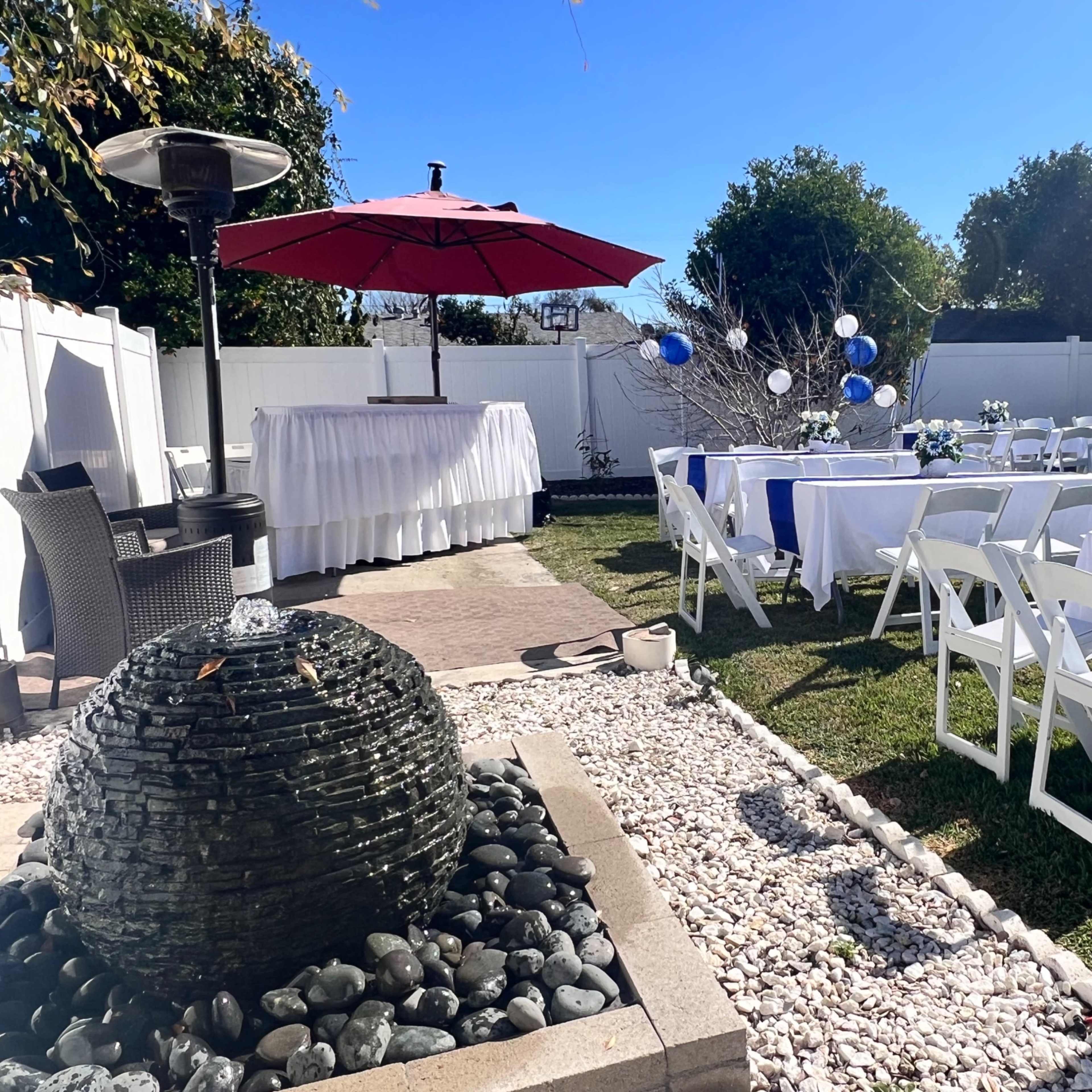 A garden party setup features white tables adorned with blue decorations, a central fountain, and an umbrella-shaded serving area.