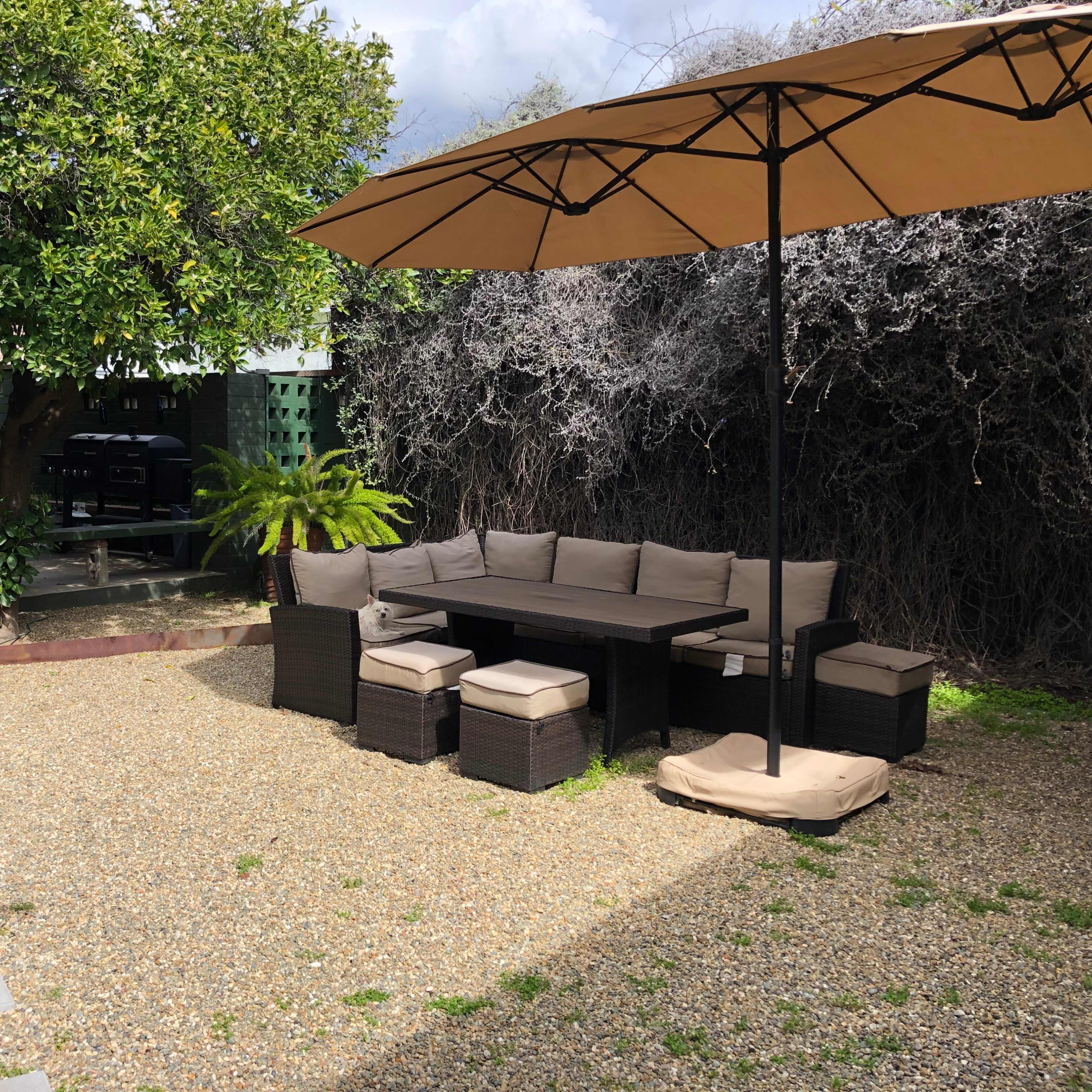 A gravel patio with a large umbrella over a sectional seating area surrounded by green foliage and a barbecue grill in the background.