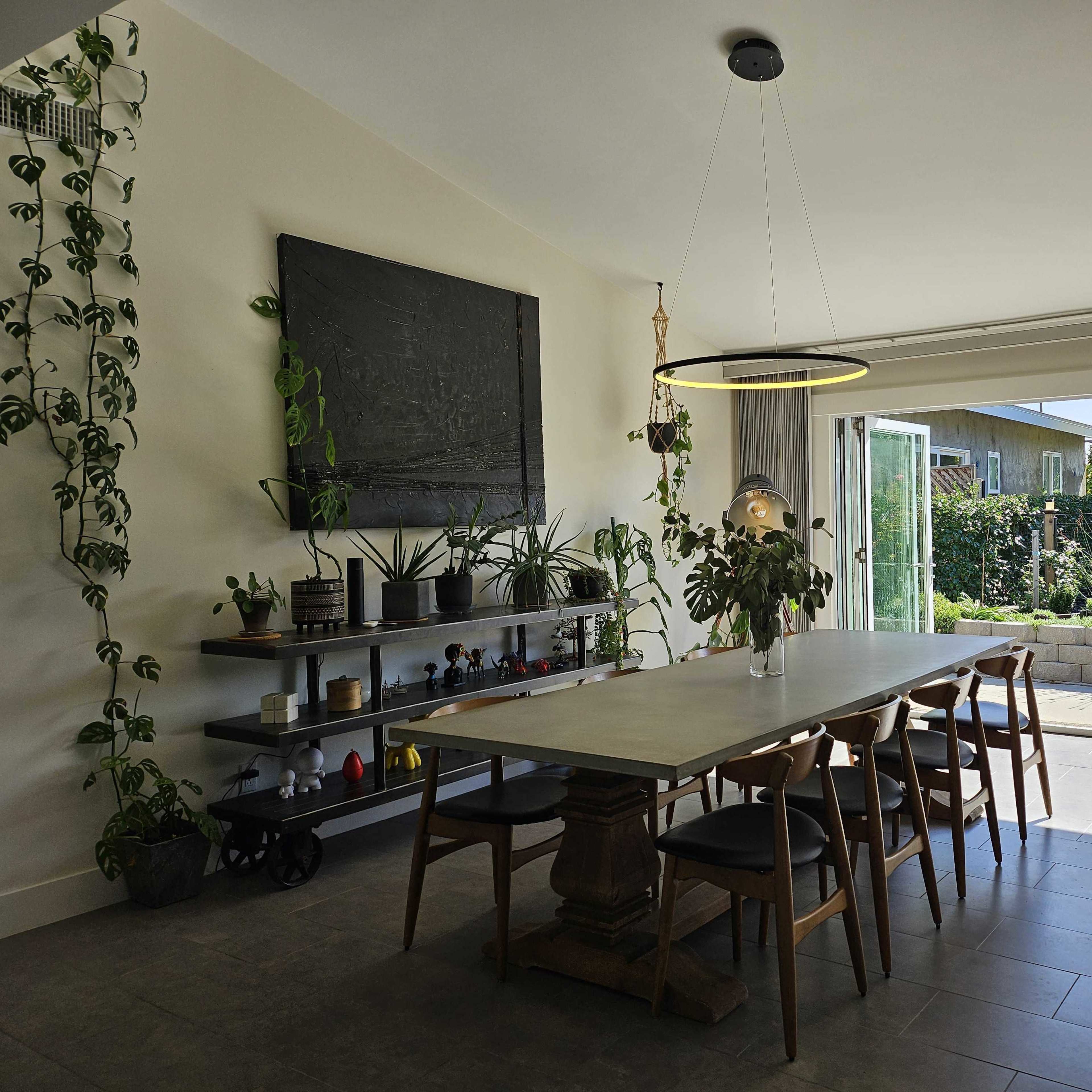 A modern dining area with a large table, surrounded by wooden chairs, and a wall adorned with plants and artwork, leading to a garden space through an open sliding door.