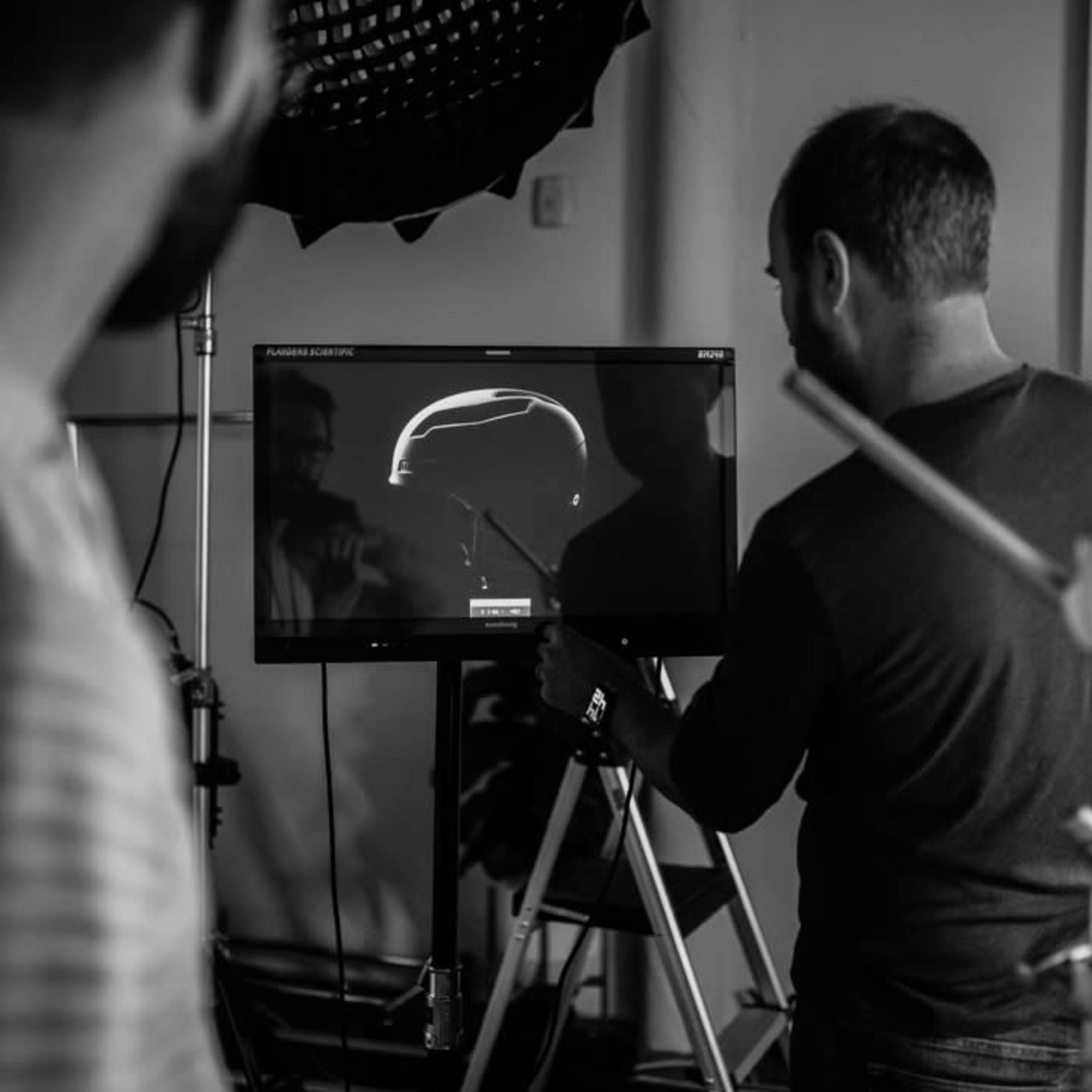A person is observing a monitor displaying a dark silhouette, while another individual stands nearby, partially visible, in a studio setting.