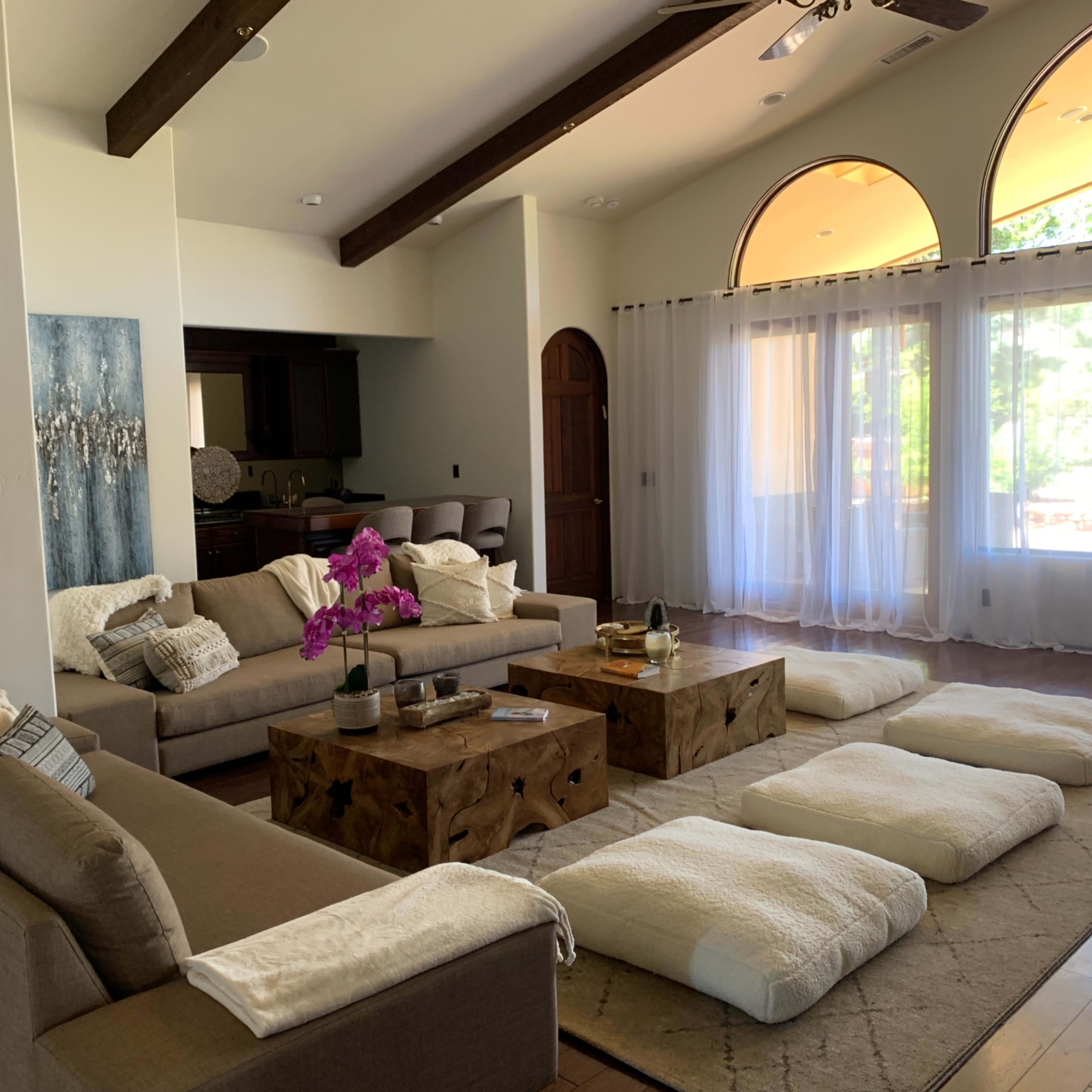 A spacious living room featuring two sofas, low wooden coffee tables, floor cushions, and large windows with natural light streaming in.