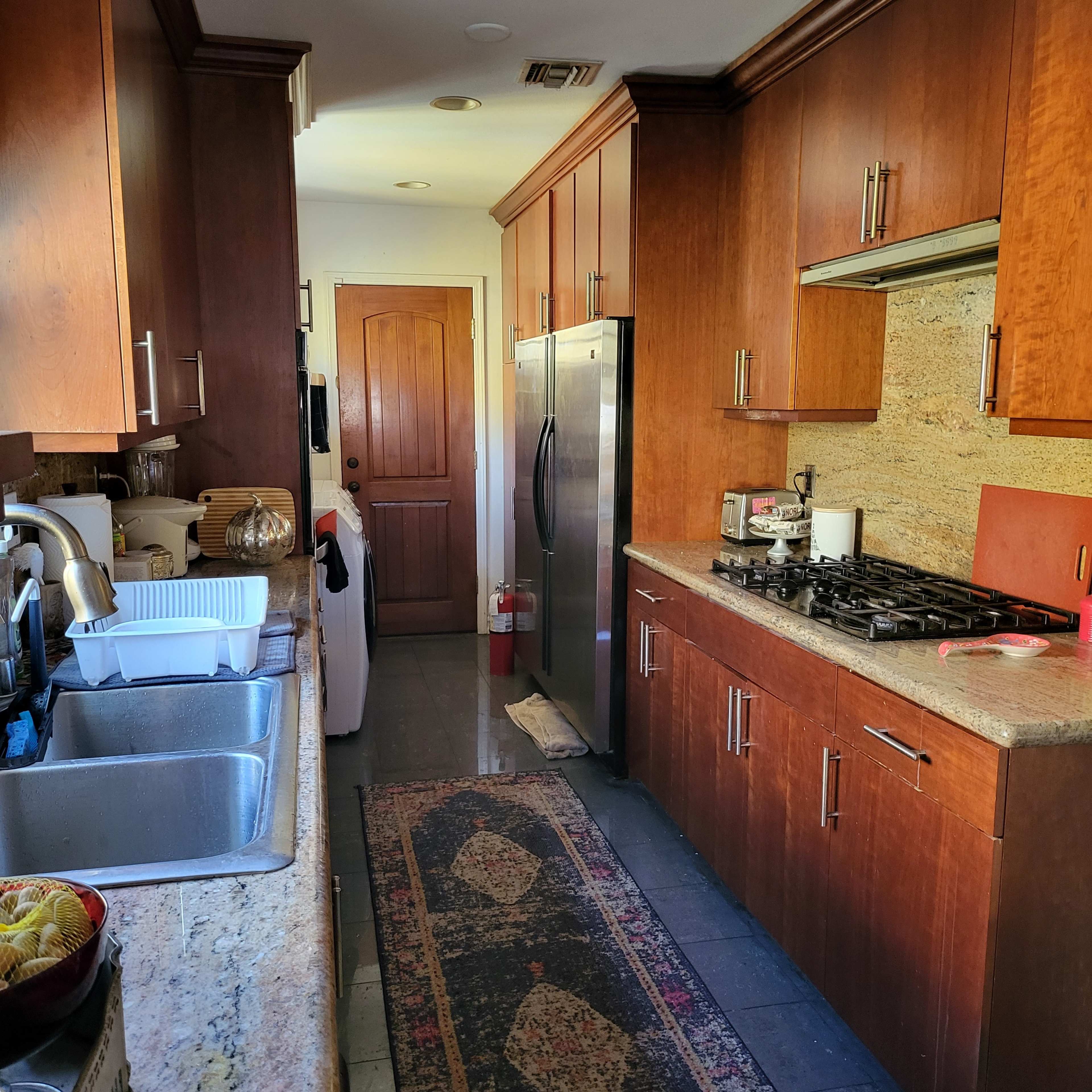A narrow kitchen features wooden cabinets, a stainless steel refrigerator, a sink, and a long countertop with a rug running the length of the floor.
