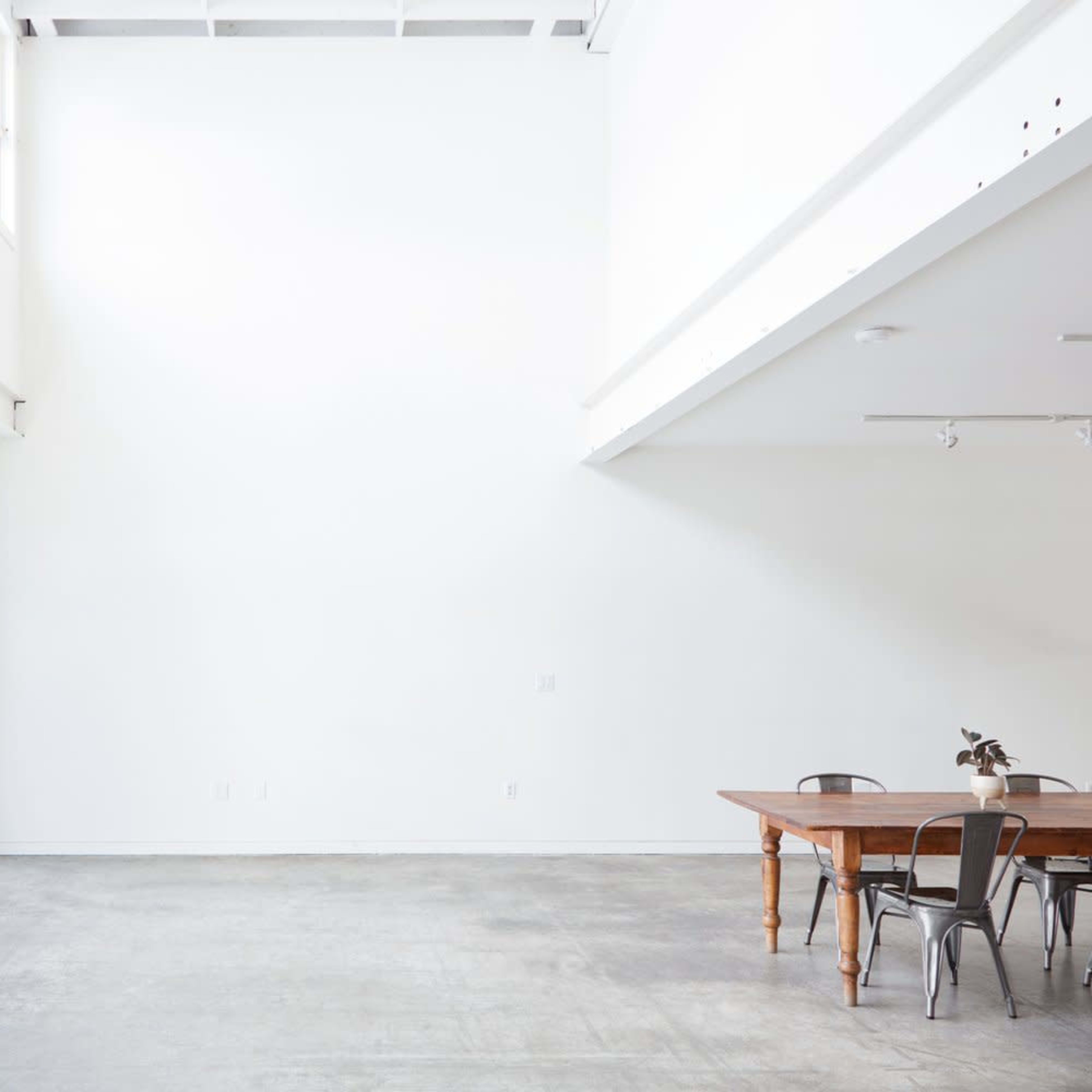 A spacious interior with high white walls, a large window, and a wooden dining table surrounded by chairs.