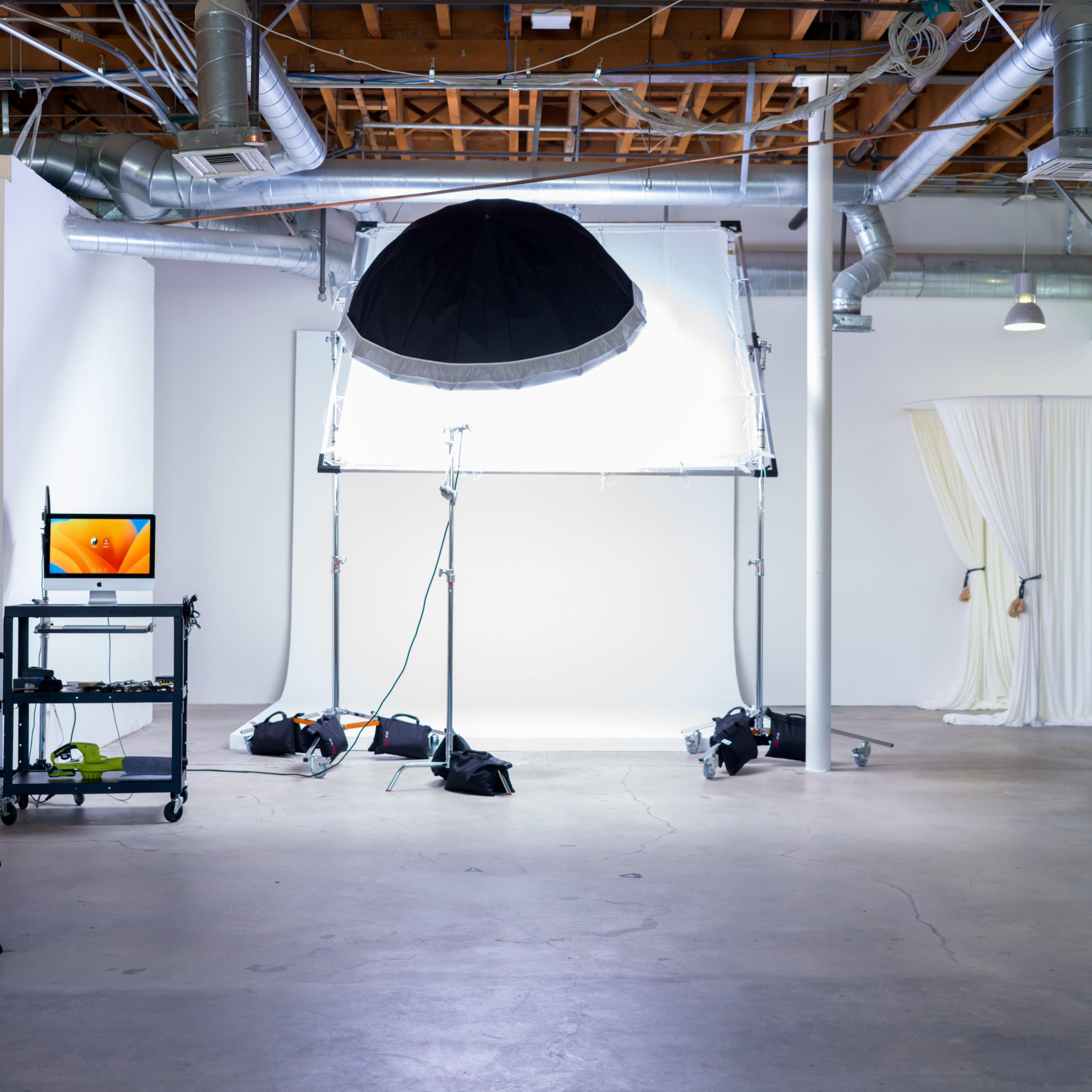 A photography studio with a large softbox light setup, a monitor on a cart, and a backdrop featuring draped fabric.