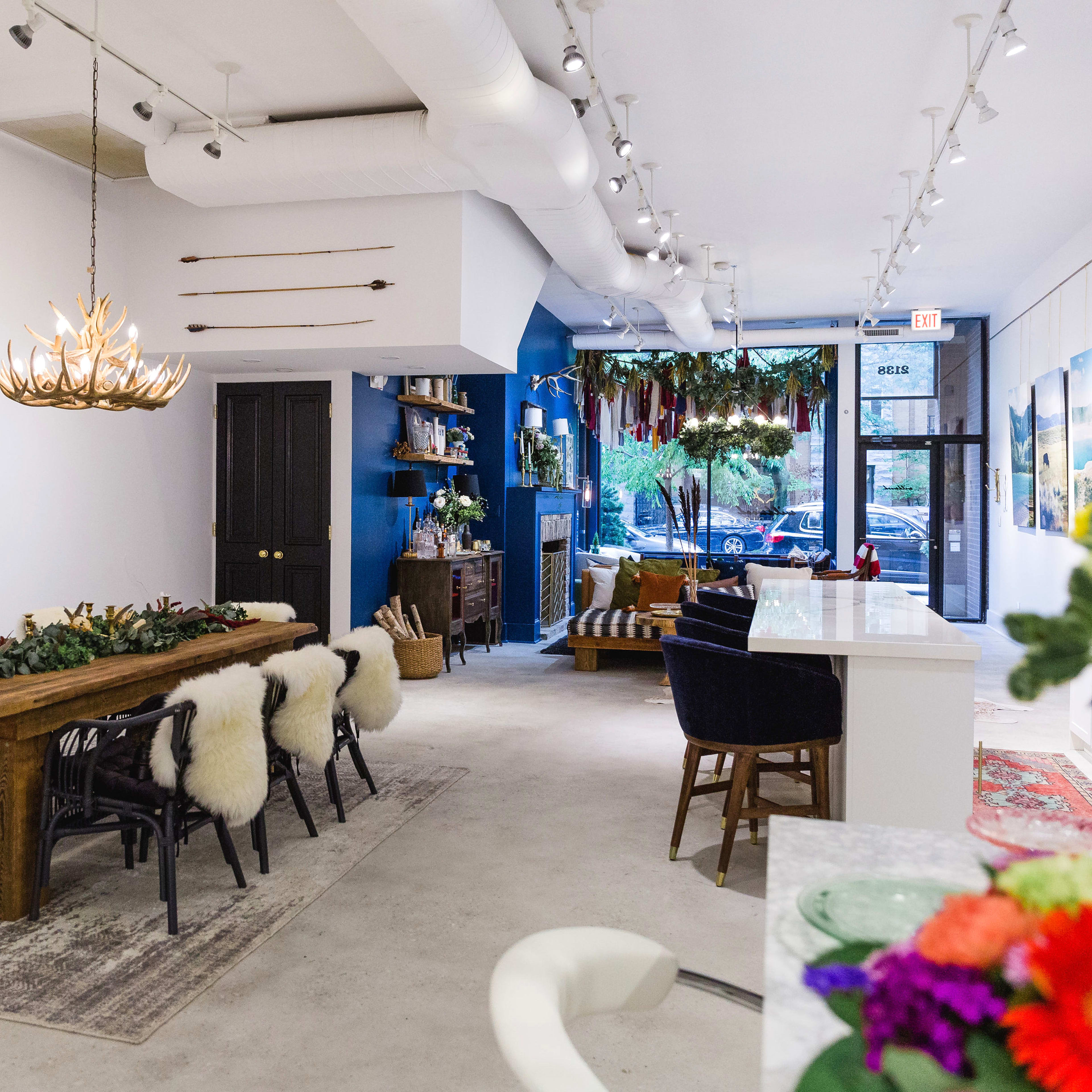 A modern interior space featuring a long wooden dining table surrounded by black chairs with white fur accents, alongside a cozy seating area and various decorative elements on the walls.