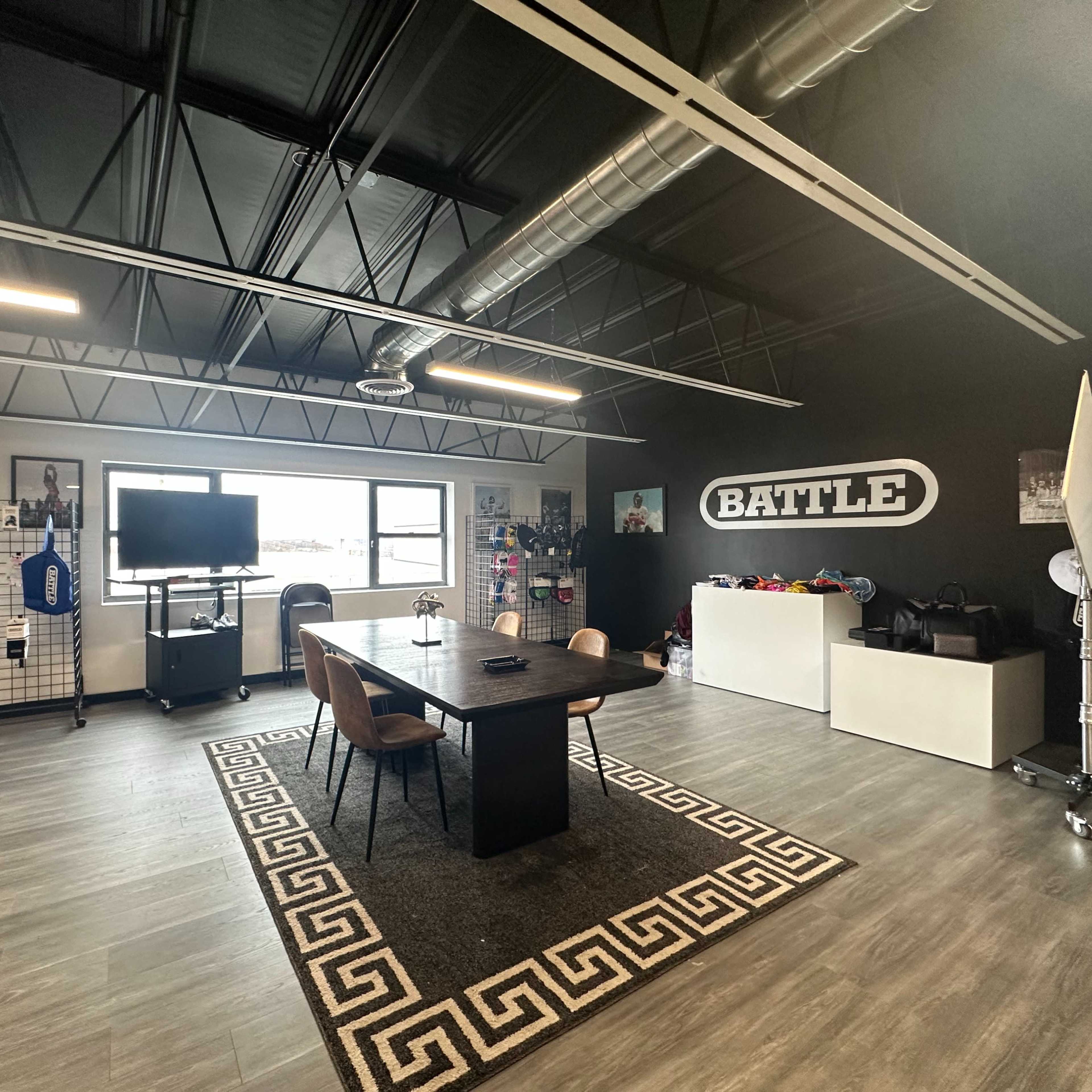 A modern office space featuring a central black table surrounded by chairs, a large screen on a stand, and various sports equipment displayed on the walls.