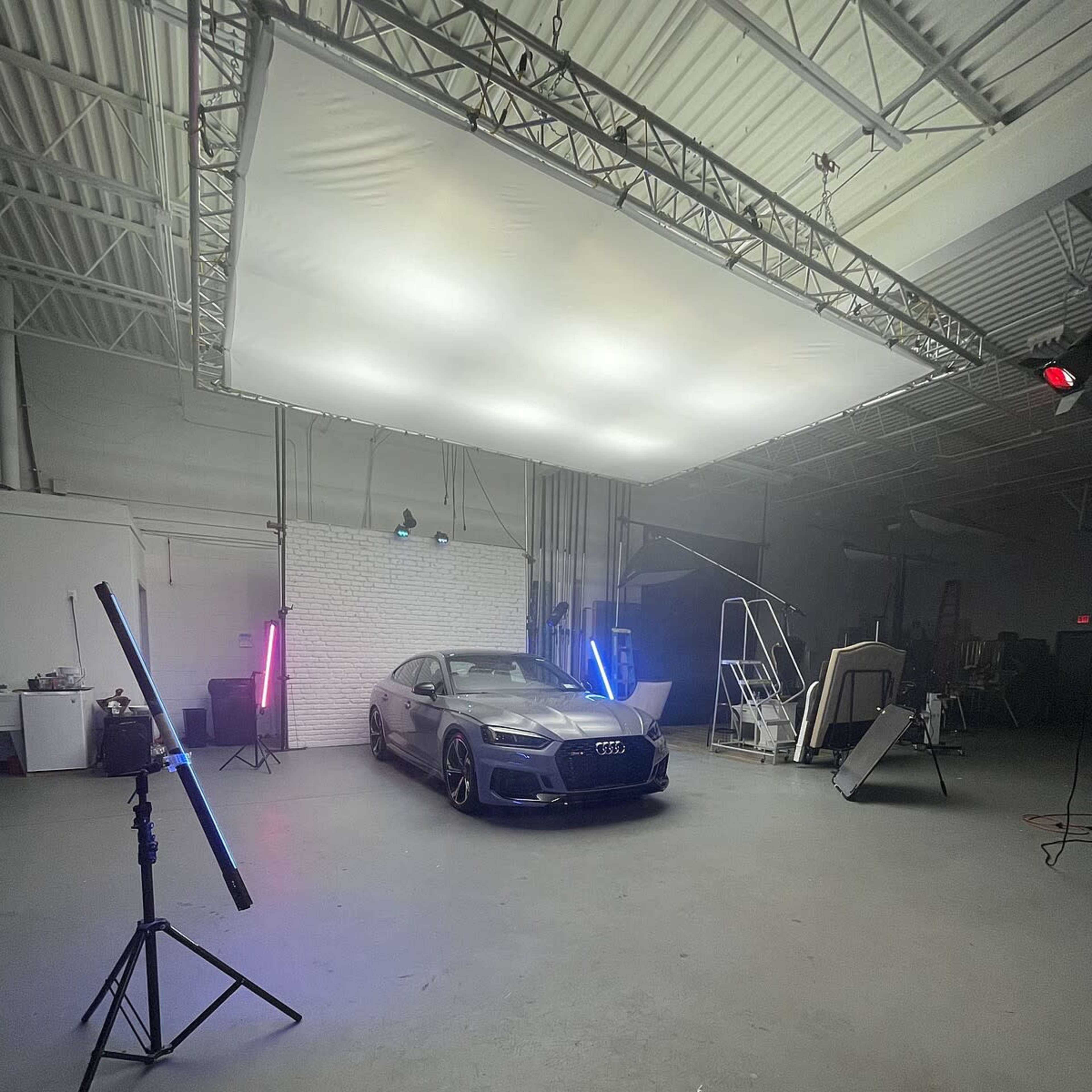 A black Audi is positioned in a studio setting with soft lighting, along with various equipment and backdrops visible.