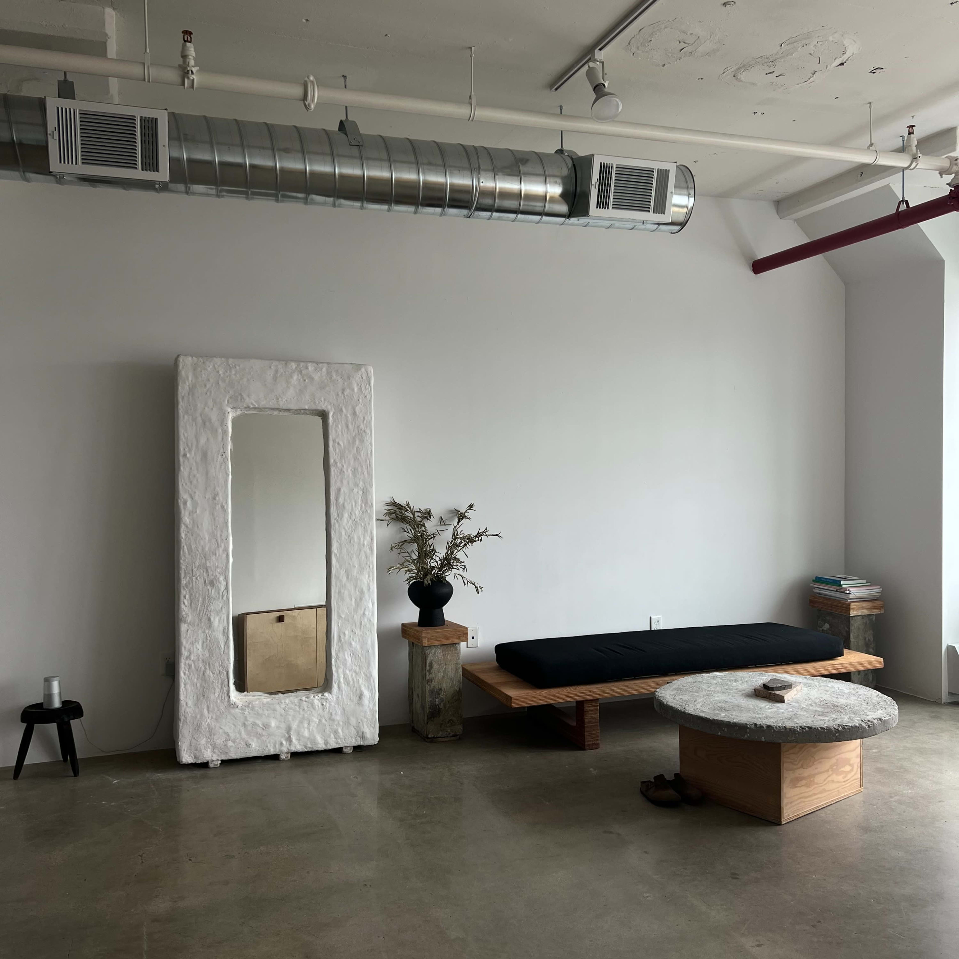 A minimalist room with a large mirror, a low wooden bed, a round coffee table, and a vase of dried flowers on a pedestal.