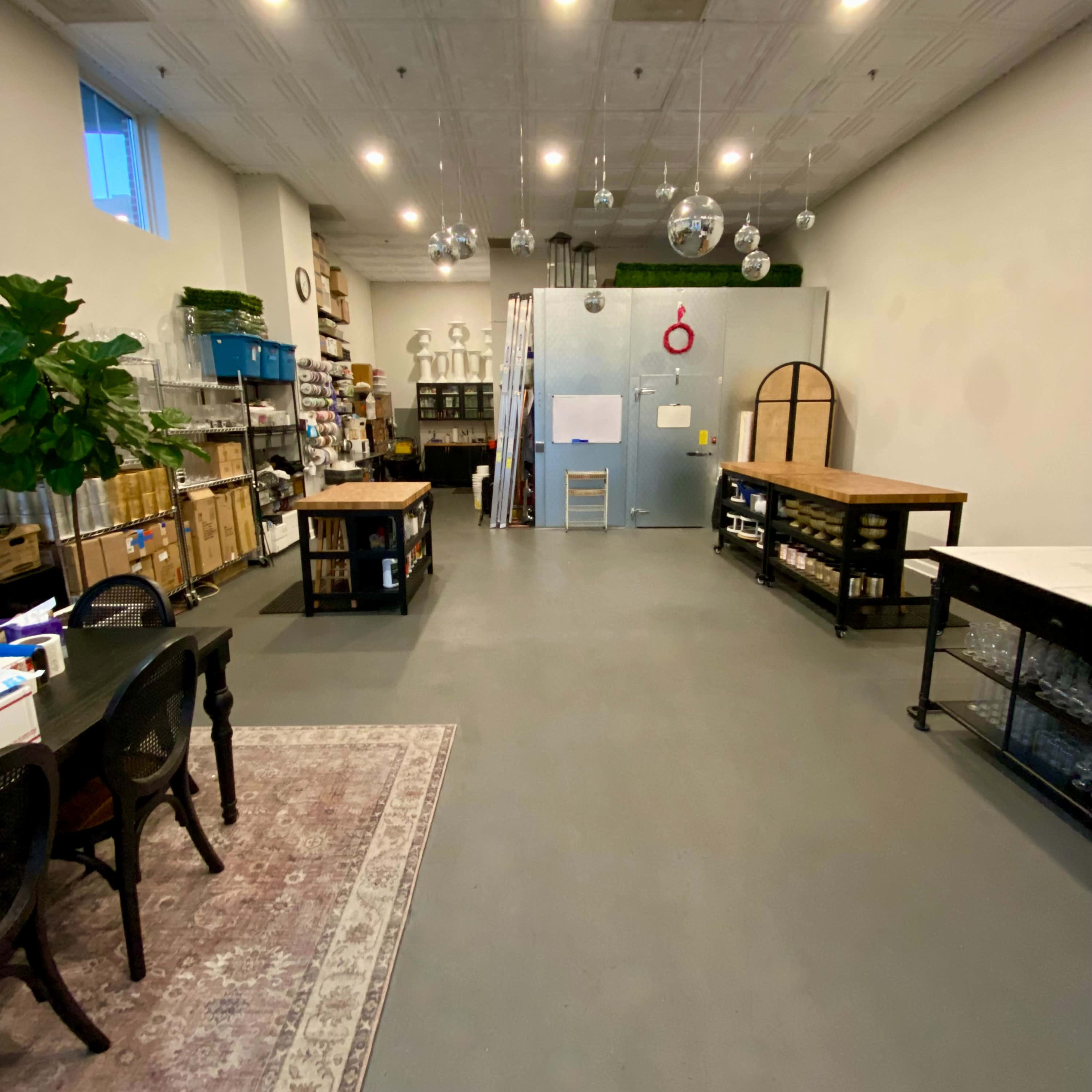 A spacious room with shelves of supplies, tables, and a large refrigerator at the back, illuminated by ceiling lights.