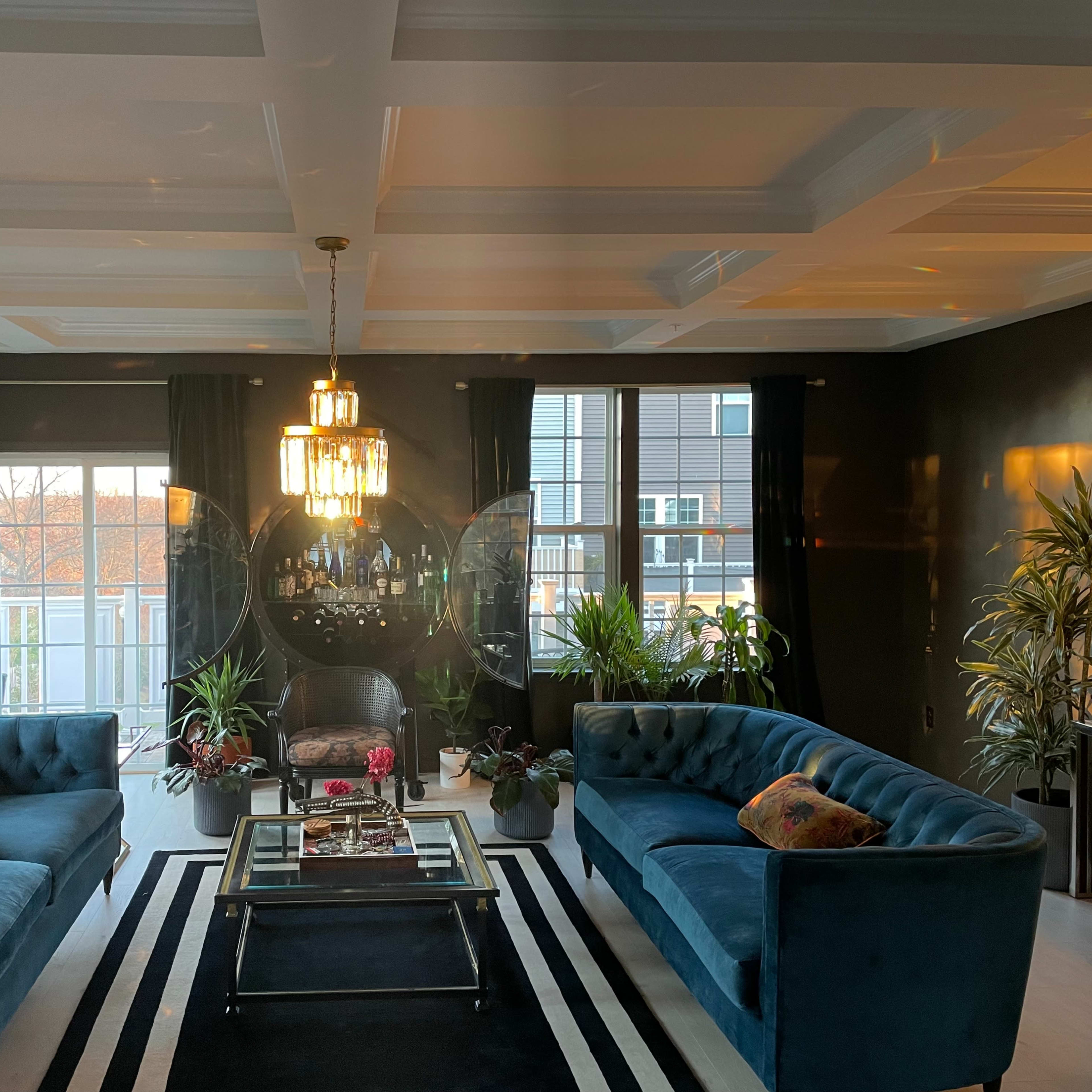 A stylish living room with two blue sofas facing each other, a coffee table in the center, and large windows surrounded by indoor plants.