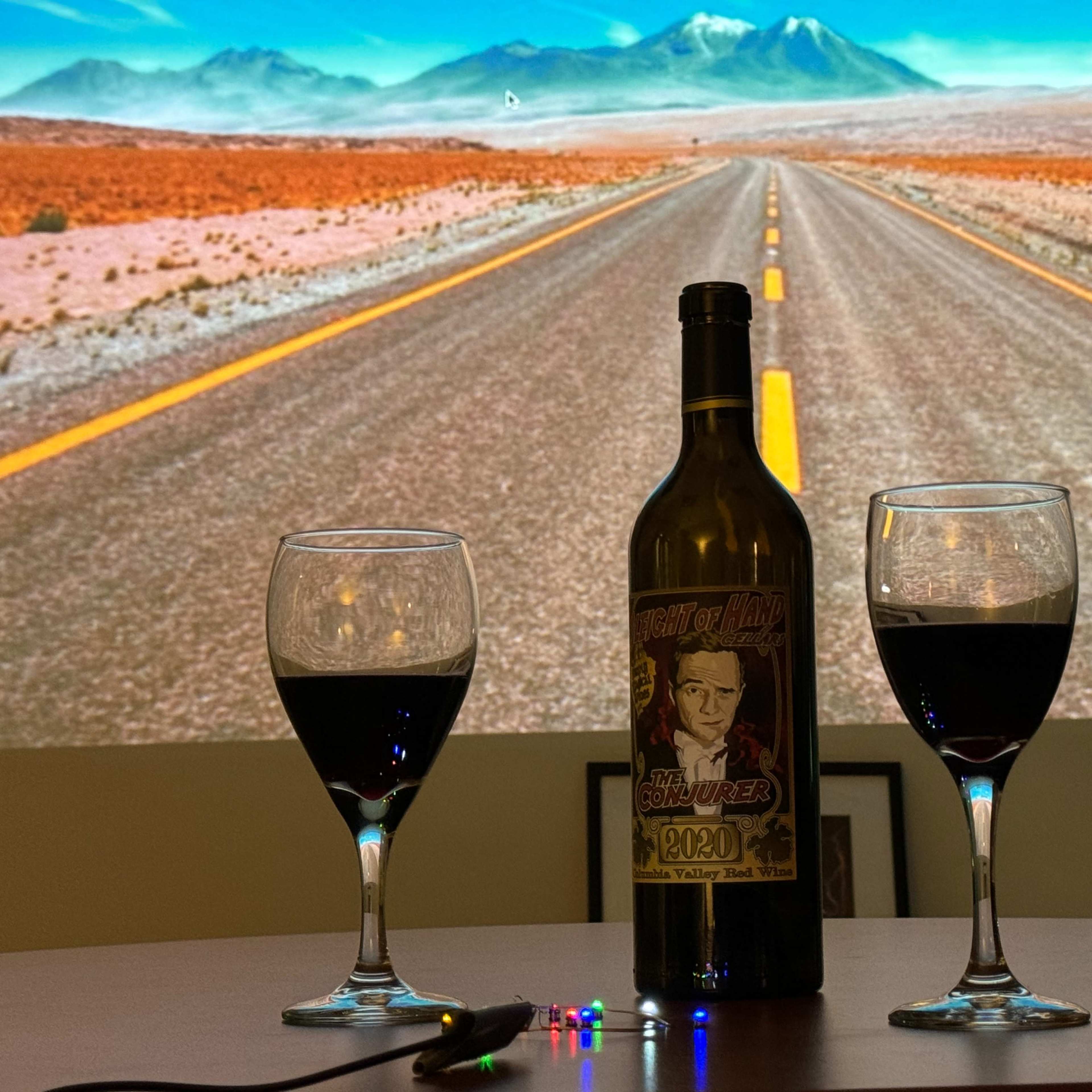 A bottle of wine and two glasses filled with red wine are positioned on a table in front of a projected image of a desert road with mountains in the background.