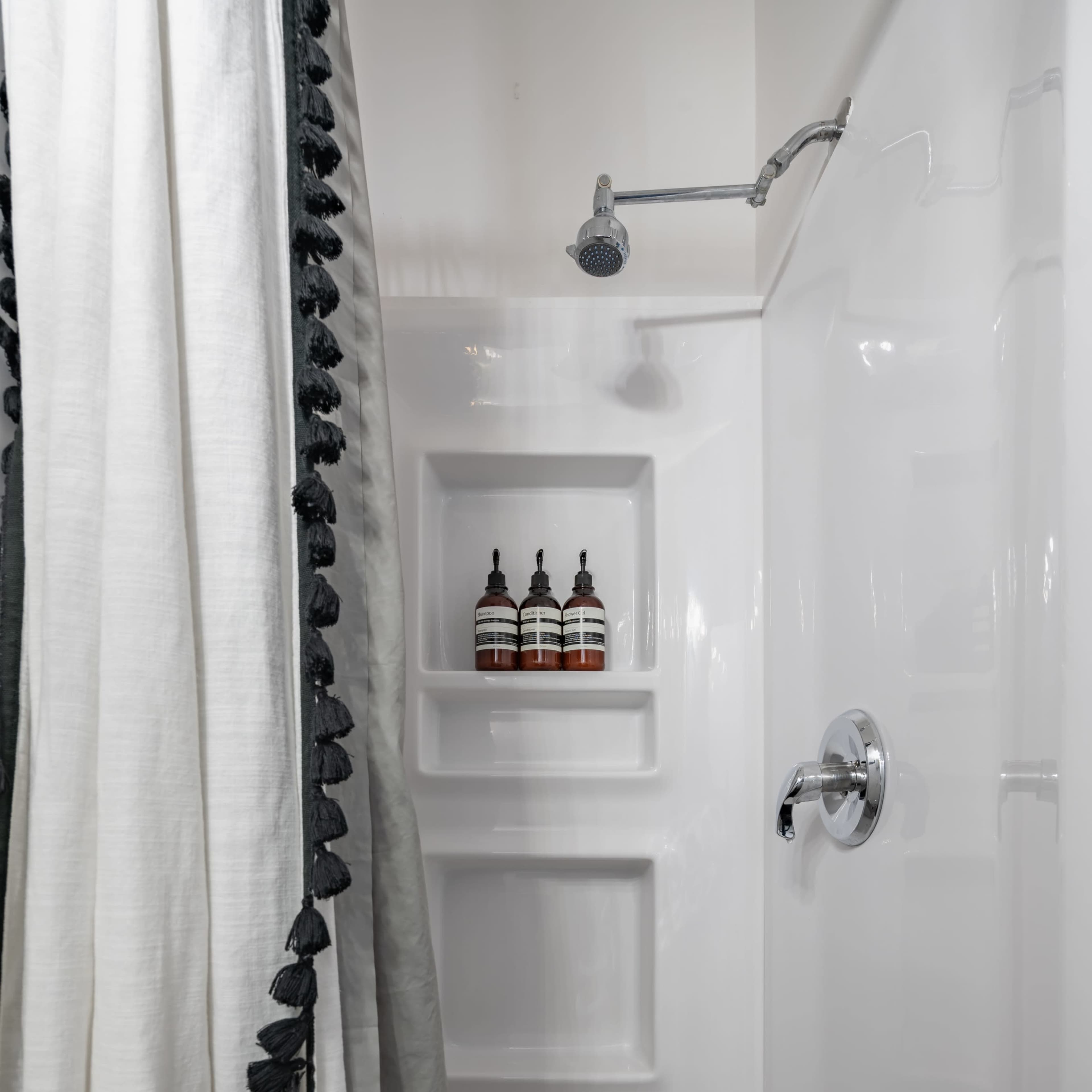 A shower area features a white tiled wall with a chrome showerhead and three bottles on a built-in shelf, partially obscured by a curtain with decorative trim.