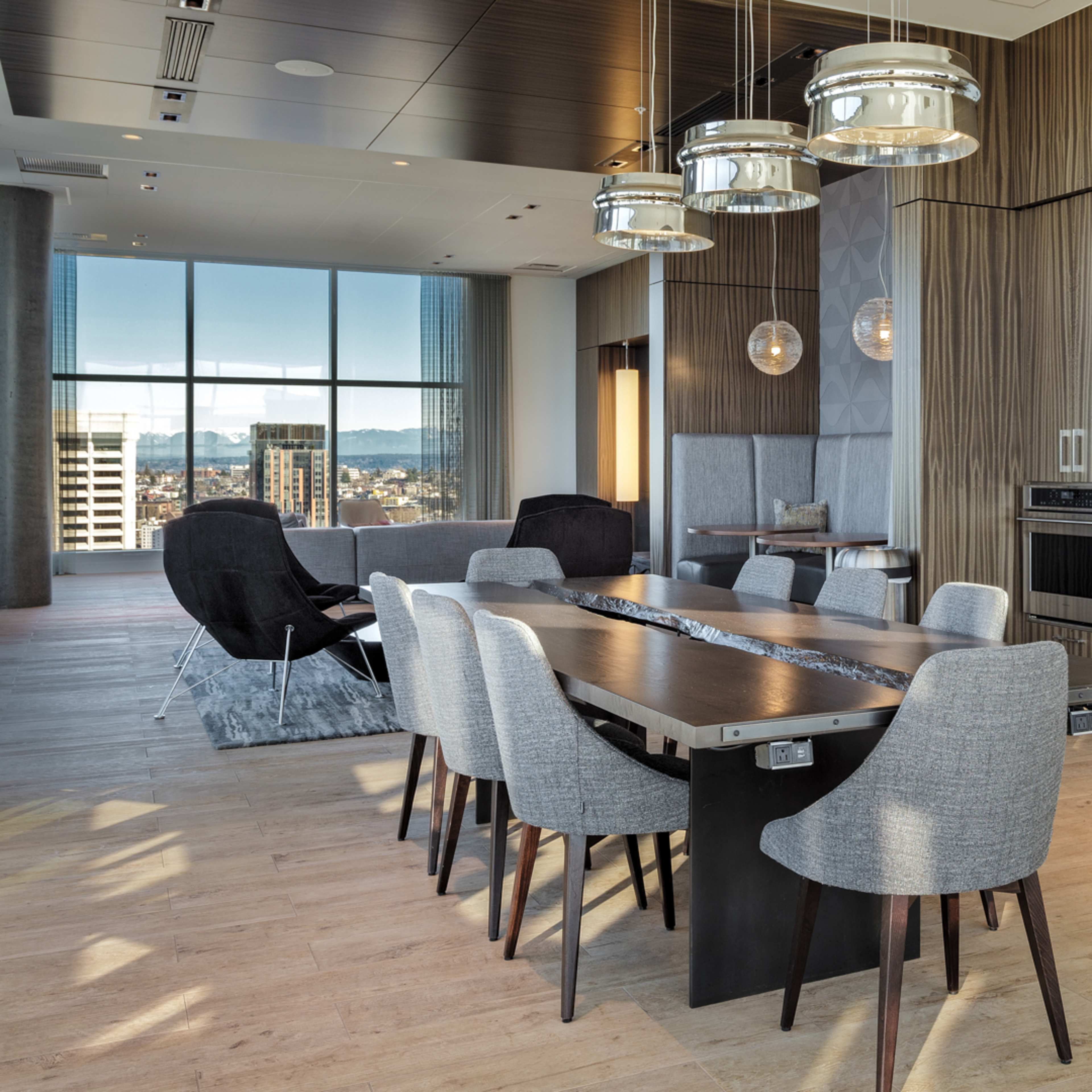 A modern dining area features a large table surrounded by gray chairs, with an adjacent kitchen equipped with stainless steel appliances and large windows offering a city view.