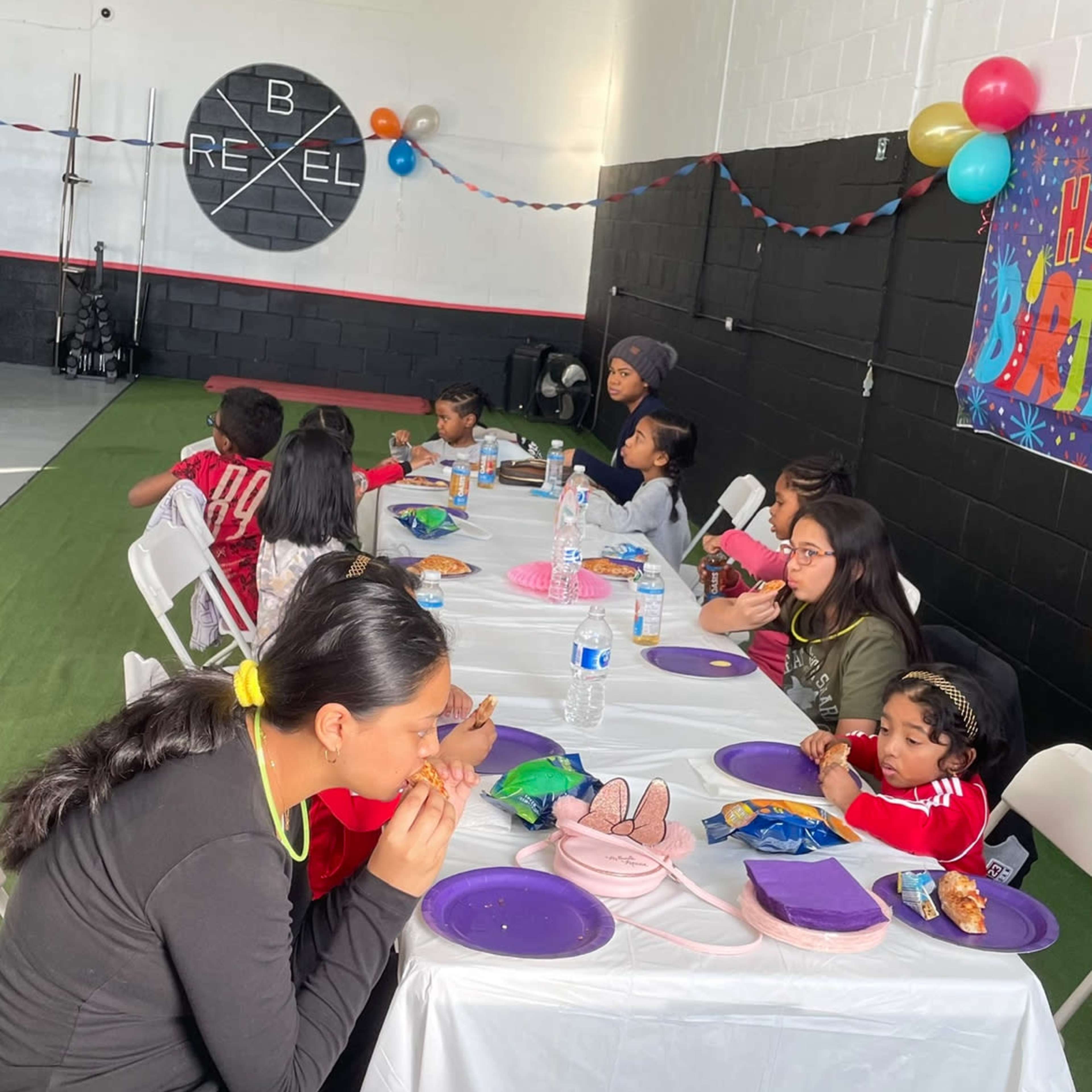 A group of children and adults sit at a long table decorated with purple plates and snacks, enjoying a celebration in a spacious indoor venue.