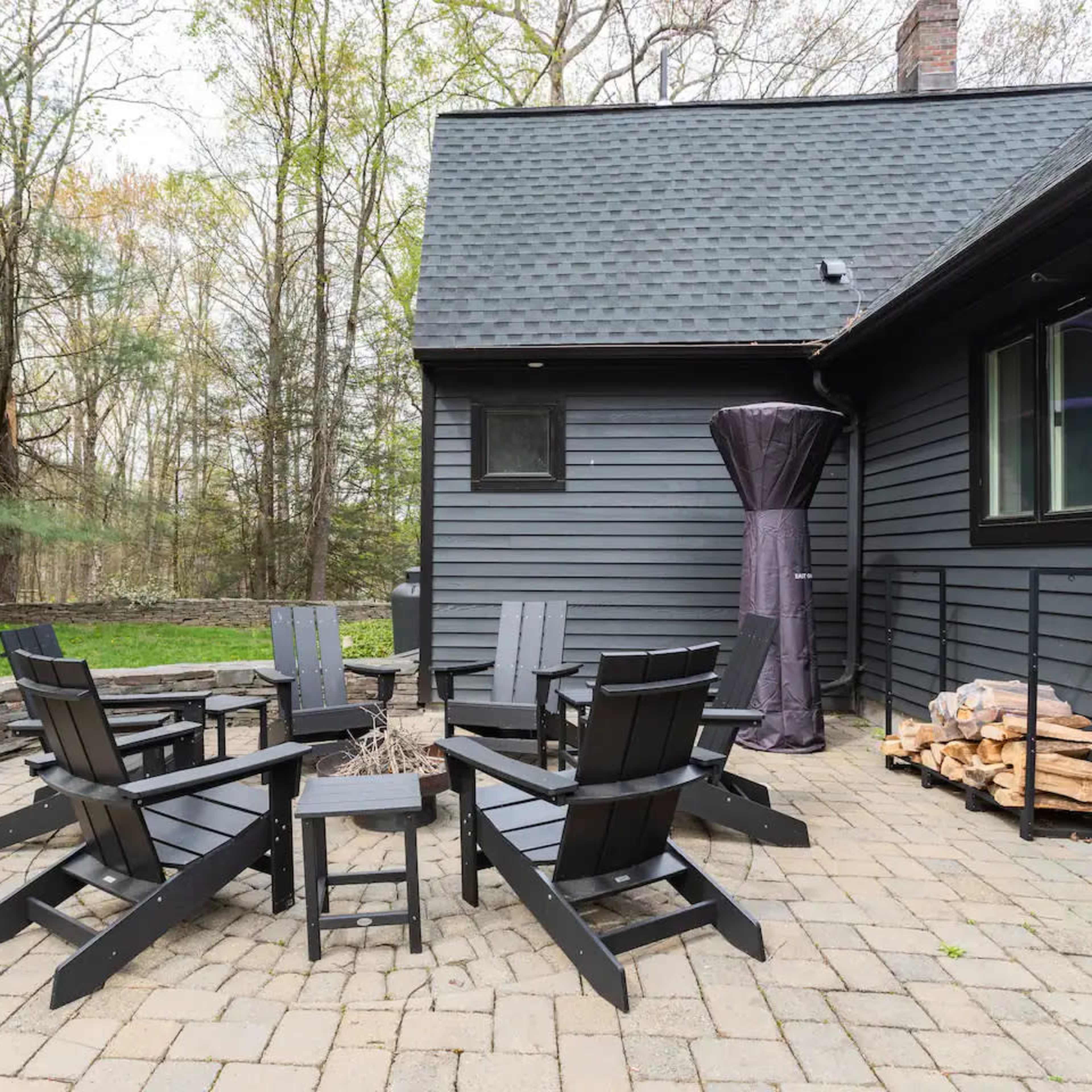 A stone patio features six black Adirondack chairs arranged around a small fire pit near a wooded area.