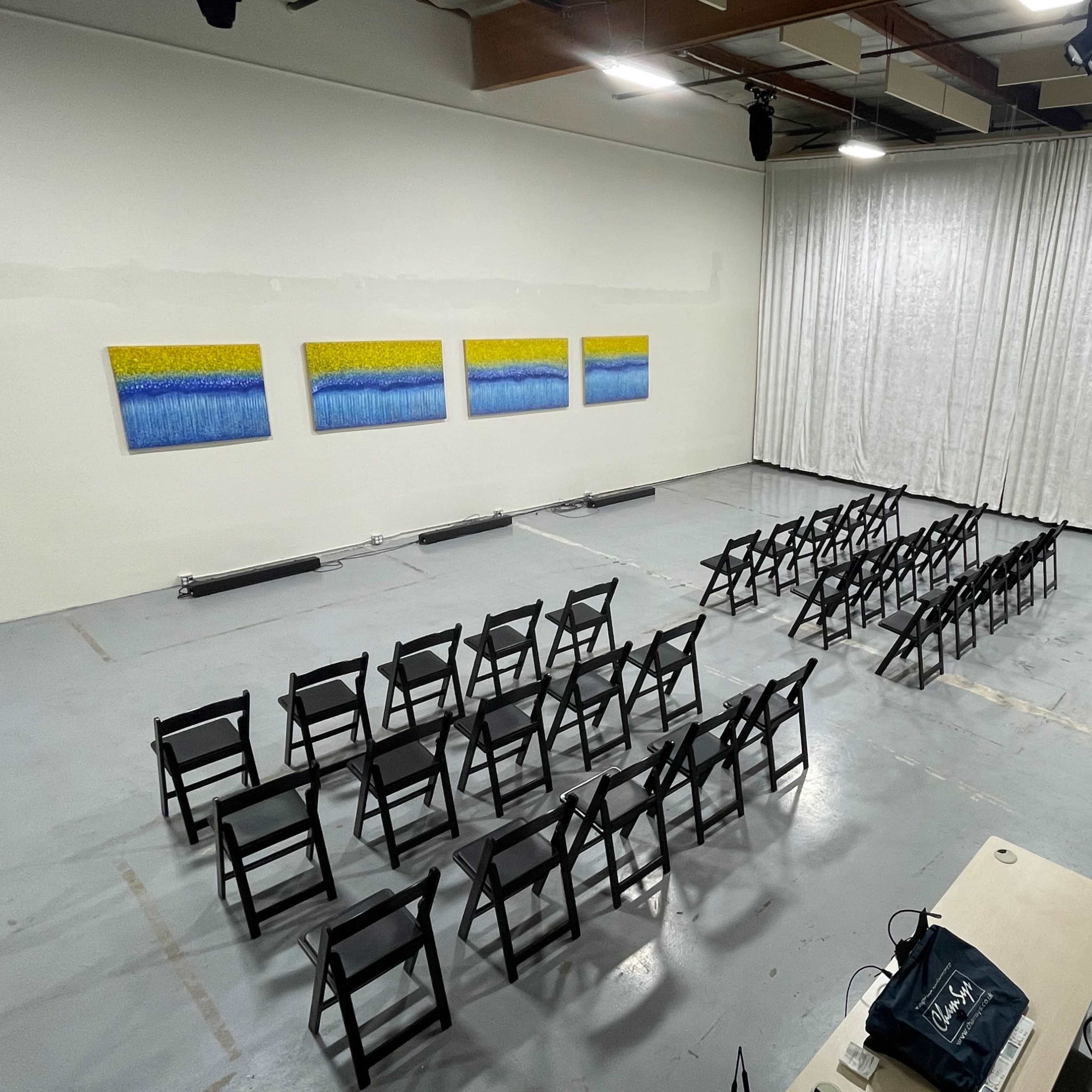 An empty room set up for an event, featuring rows of black chairs facing a set of three colorful paintings on the wall, with white curtains draped across one side.
