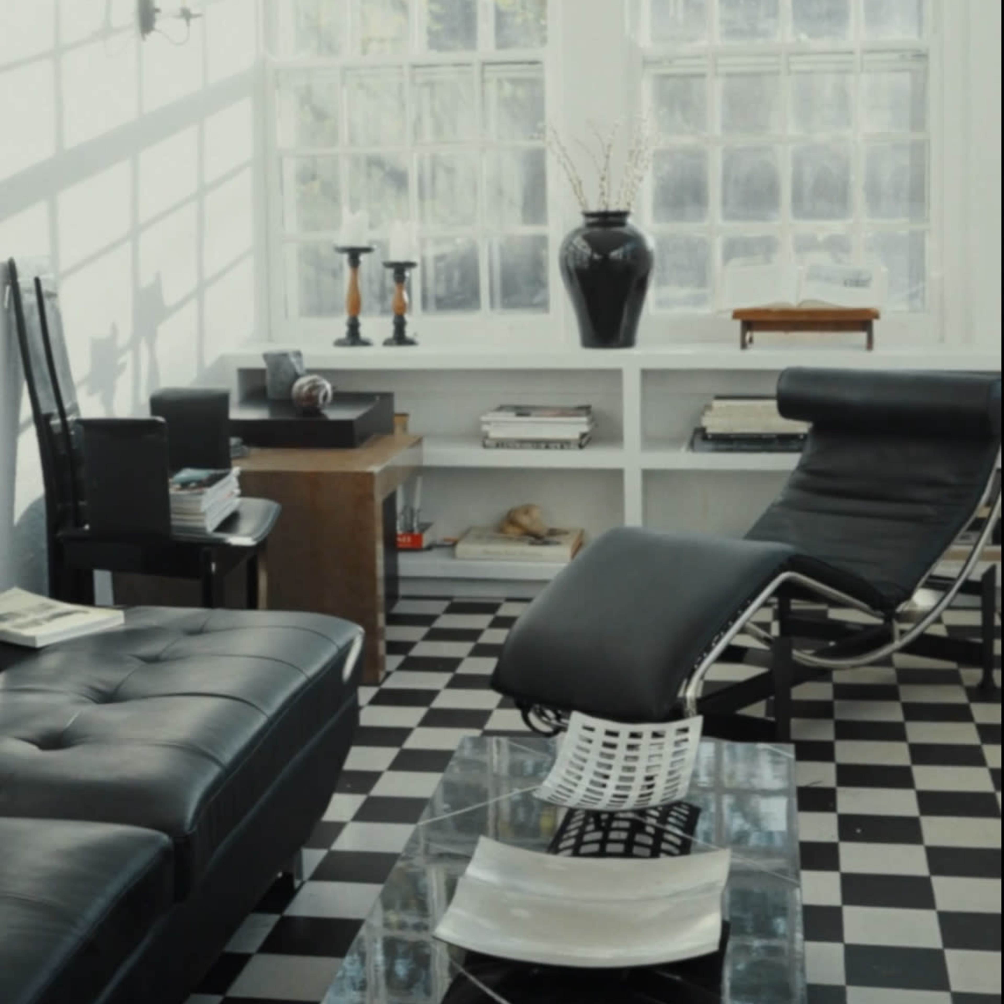 A modern living room with a black and white checkered floor, a black leather sofa, and a chaise lounge, illuminated by light filtering through large windows.