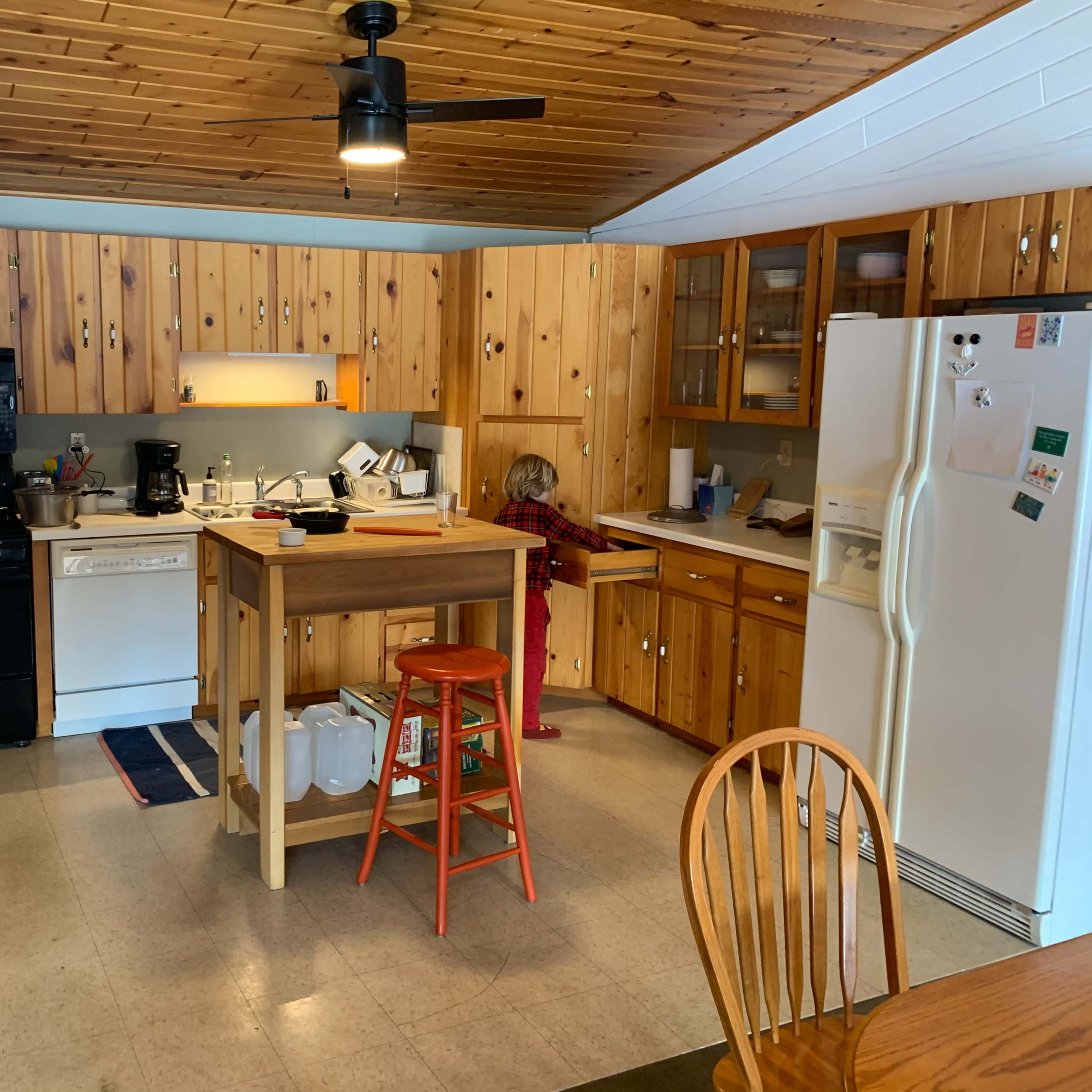 A kitchen features wooden cabinetry, a central island with a stool, and various appliances, including a stove, dishwasher, and refrigerator.