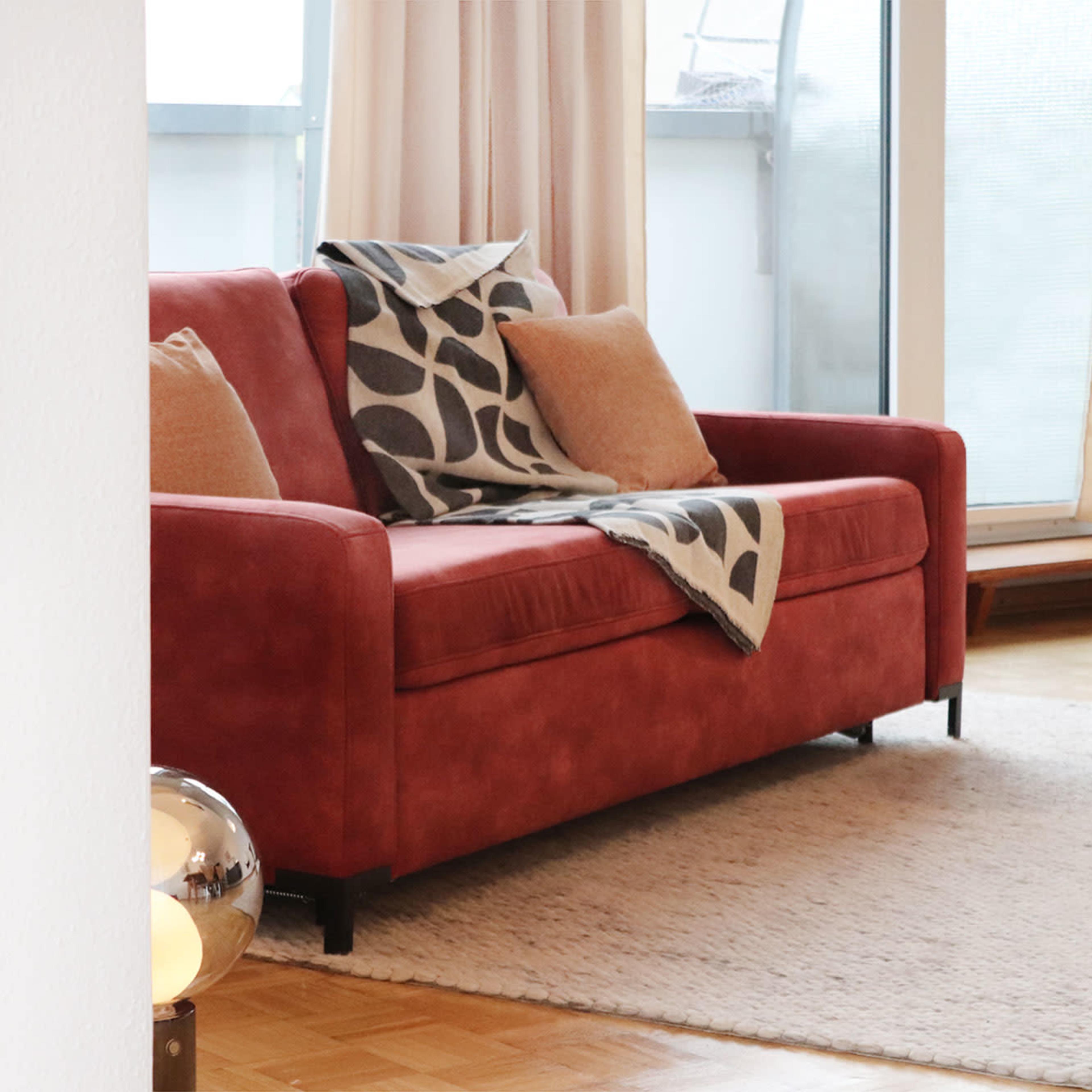 A red couch with pillows and a patterned throw is positioned in a bright living room near a textured rug and a wooden side table.
