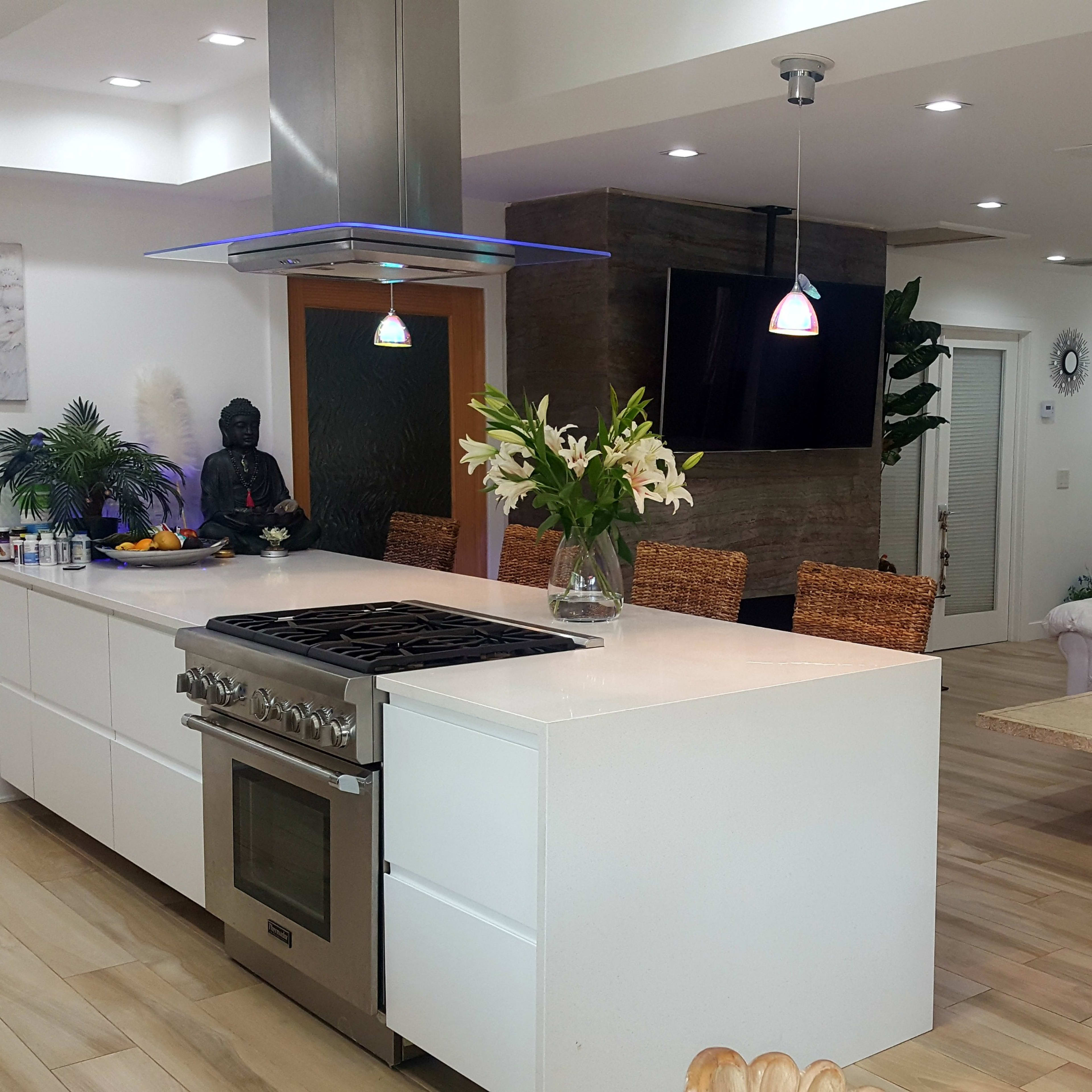 A modern kitchen with a large island, stainless steel appliances, and a dining area featuring wicker chairs and tropical plants.