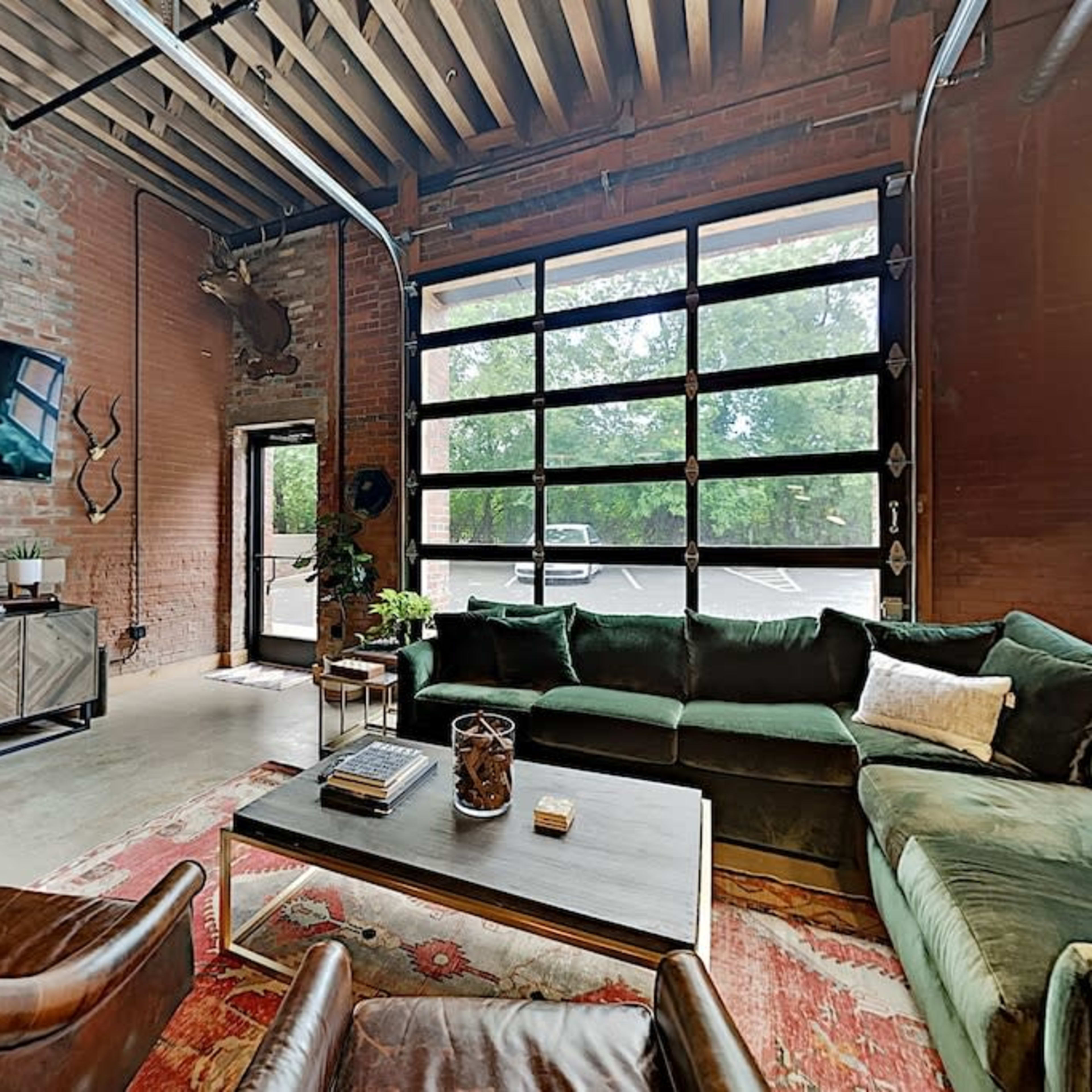A spacious living area with exposed brick walls, large windows, and a green sectional sofa facing a coffee table.