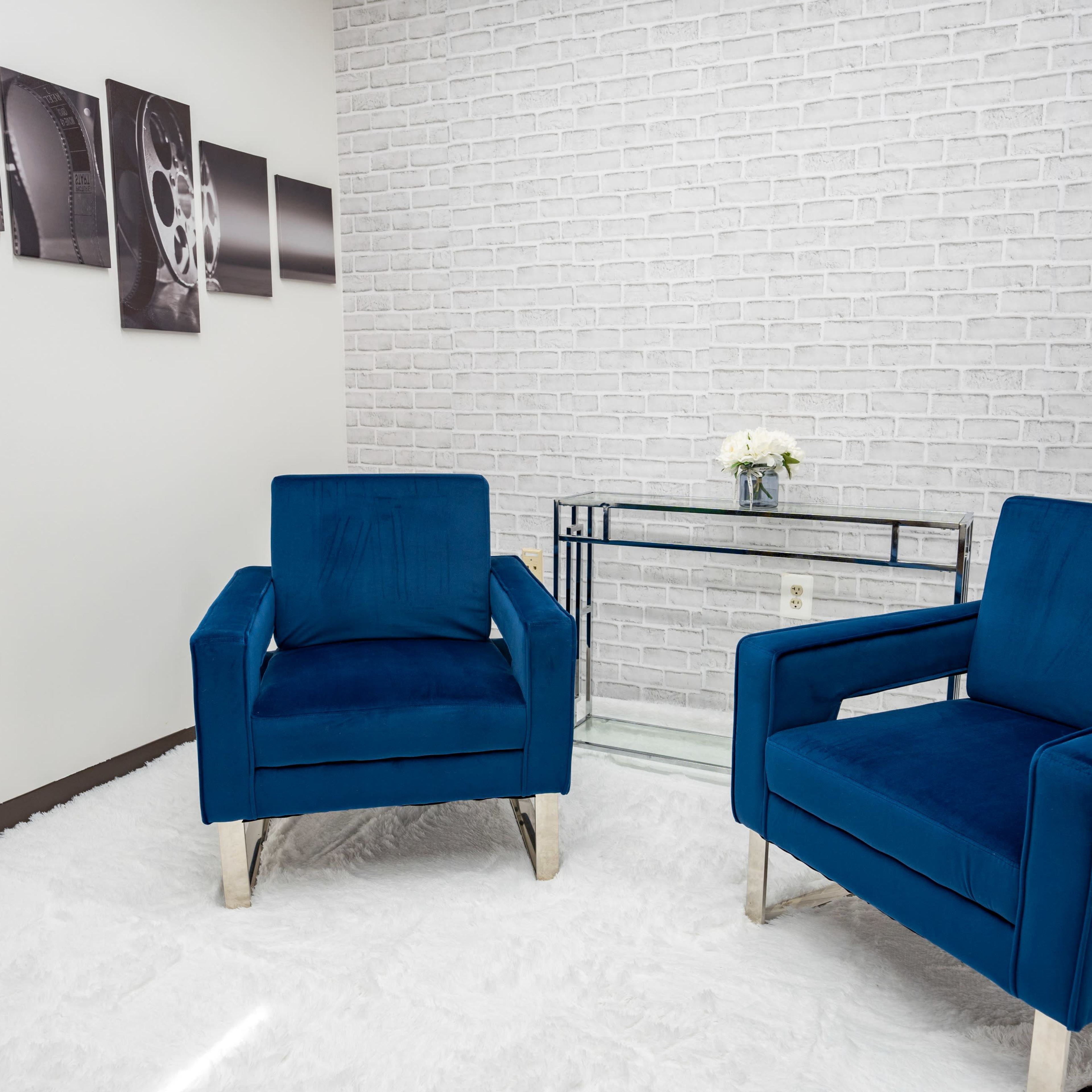 A modern sitting area with two blue chairs facing a glass table, situated against a white brick wall.
