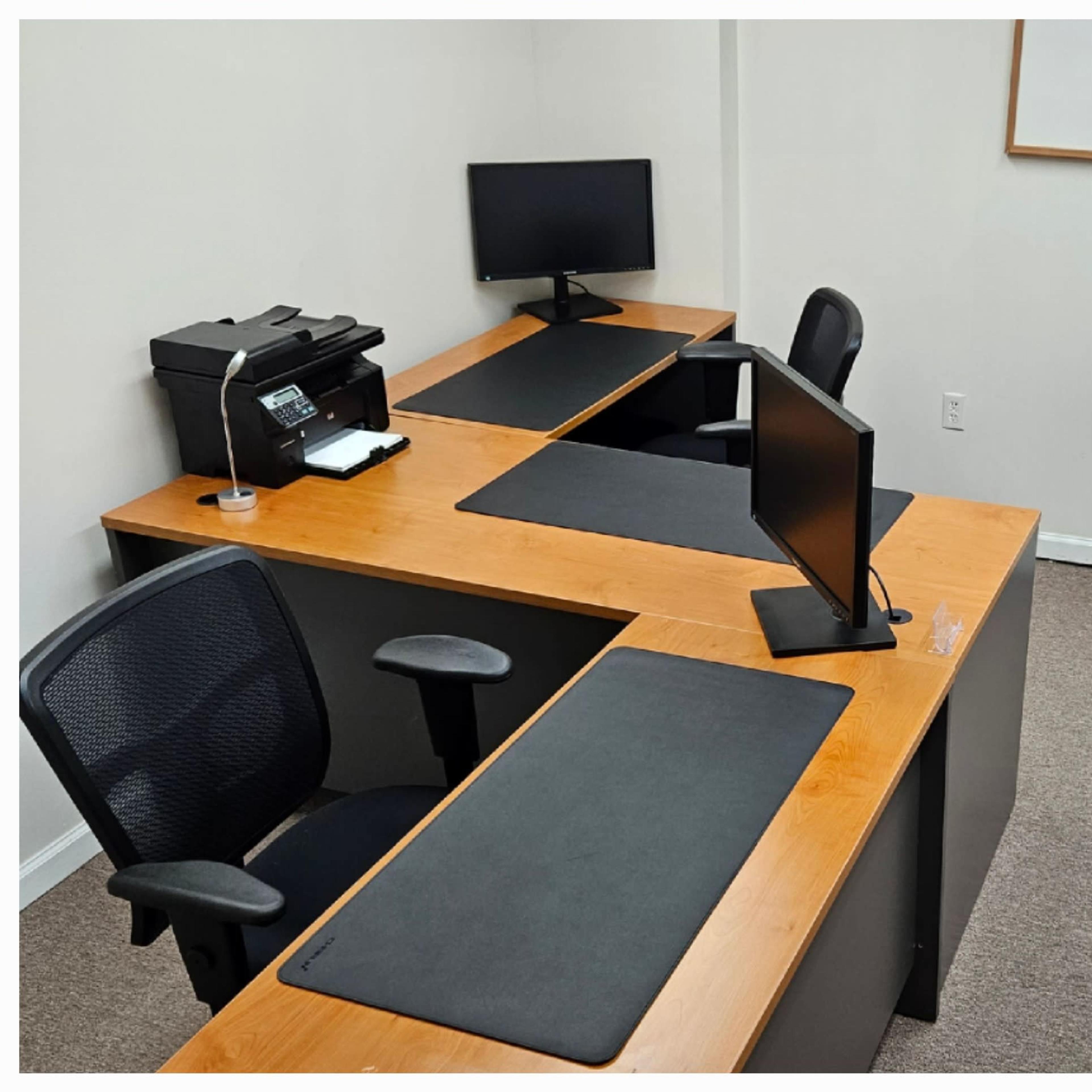 A small office space featuring two desks with computer monitors, a printer, and a heating unit along the wall.