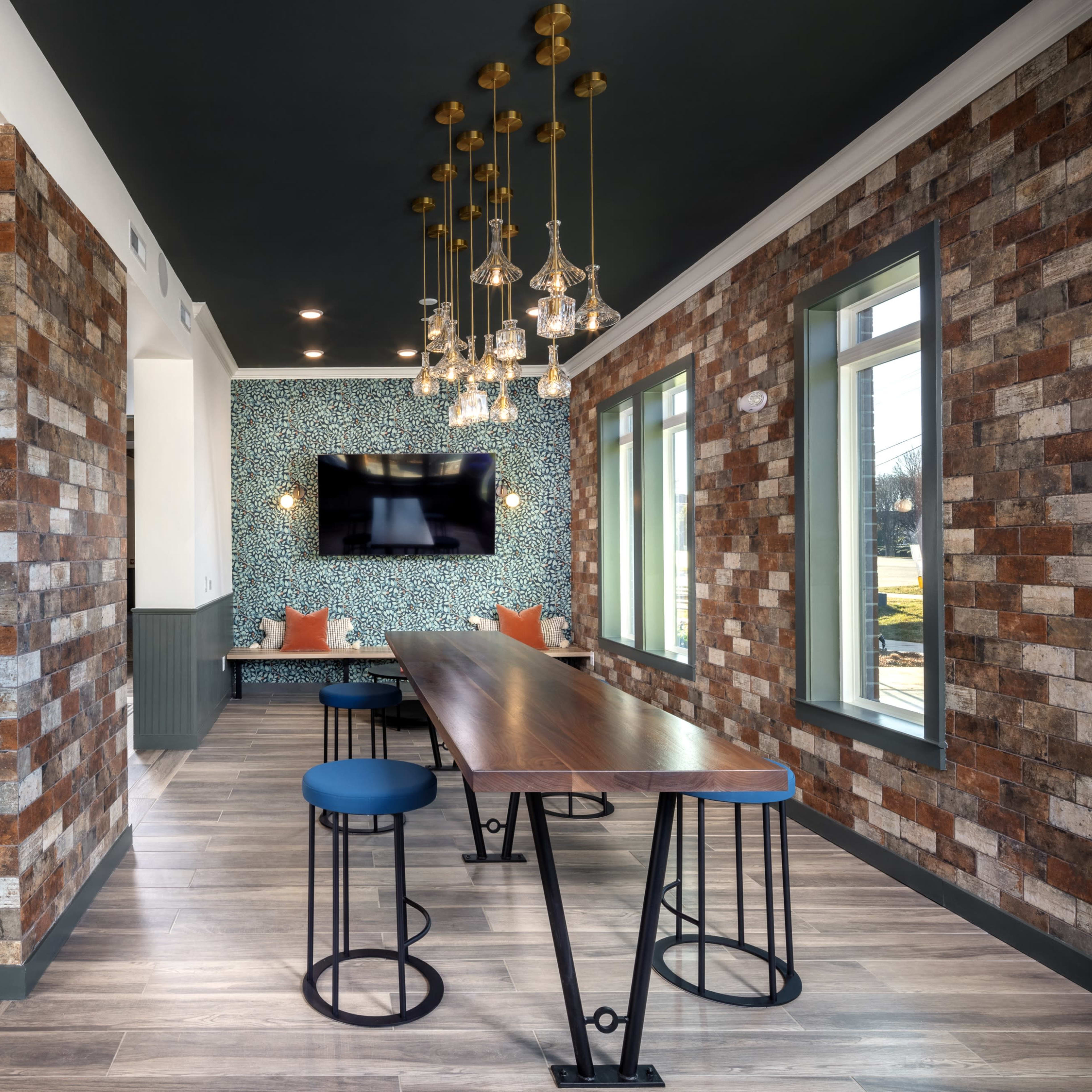 A modern communal space featuring a long wooden table with high stools, surrounded by patterned walls, a kitchen area, and decorative lighting fixtures.