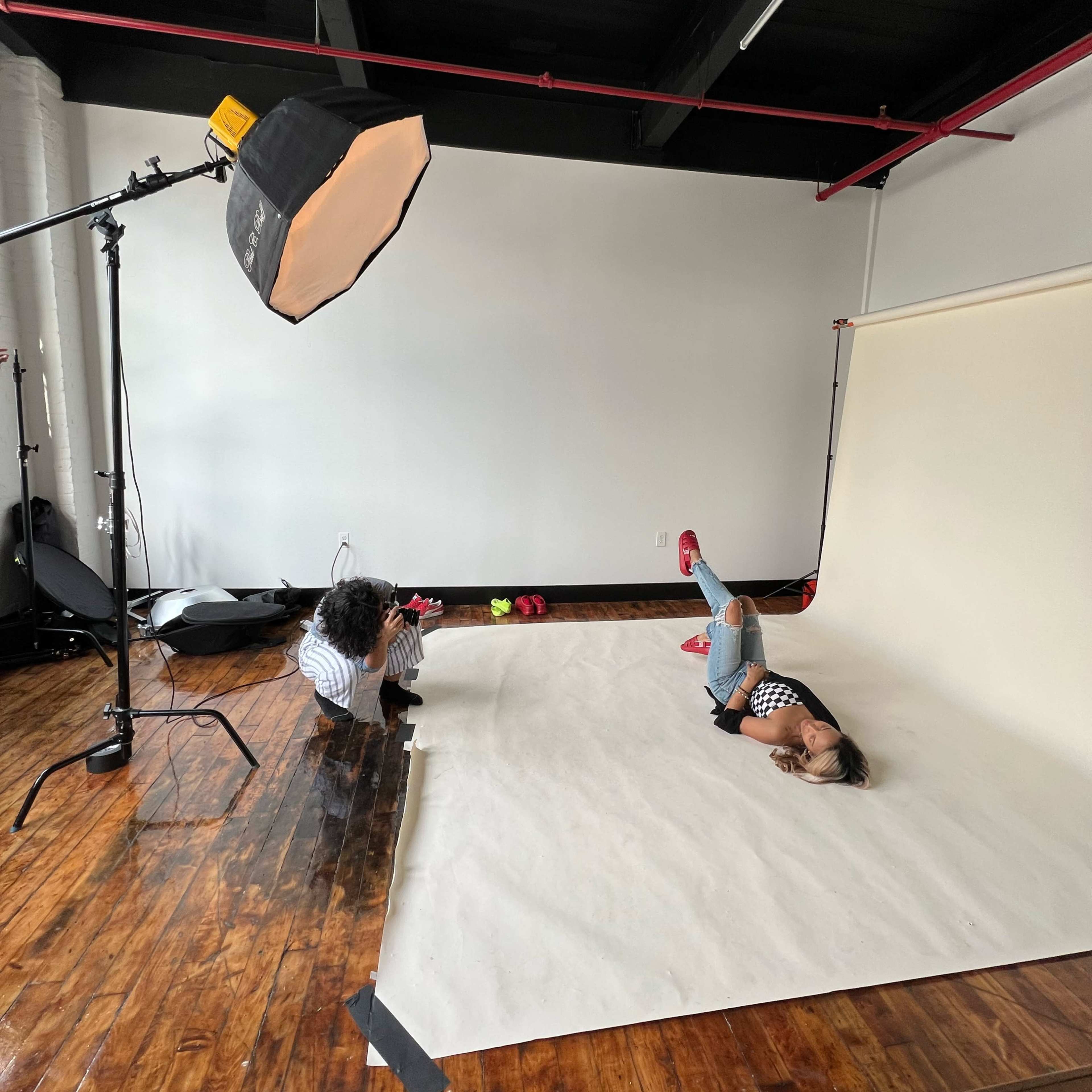 A photographer captures a model lying on a backdrop in a studio setting with lighting equipment set up nearby.