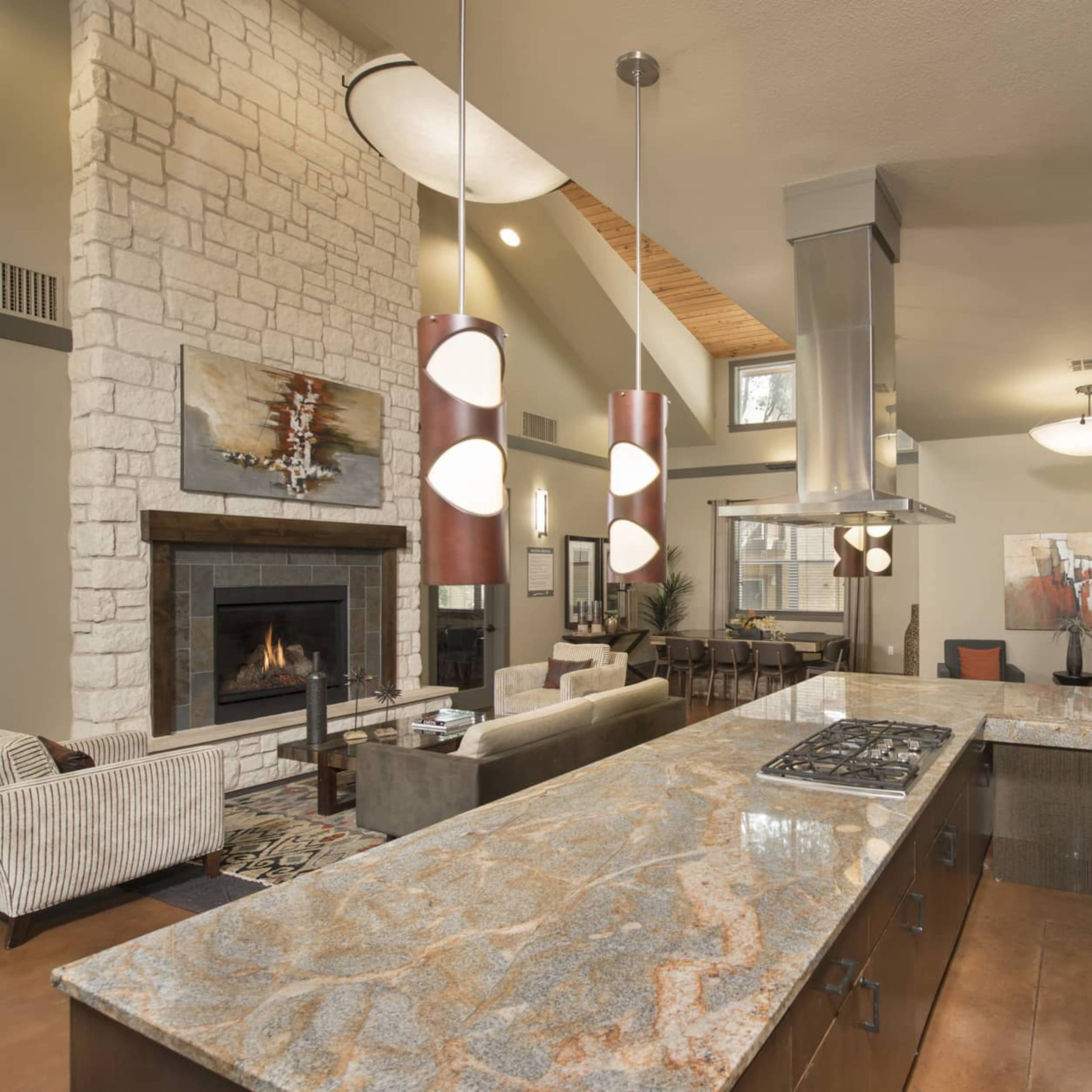 A modern kitchen connected to a living area, featuring a large island with a marble countertop, stainless steel appliances, and a stone fireplace in the background.