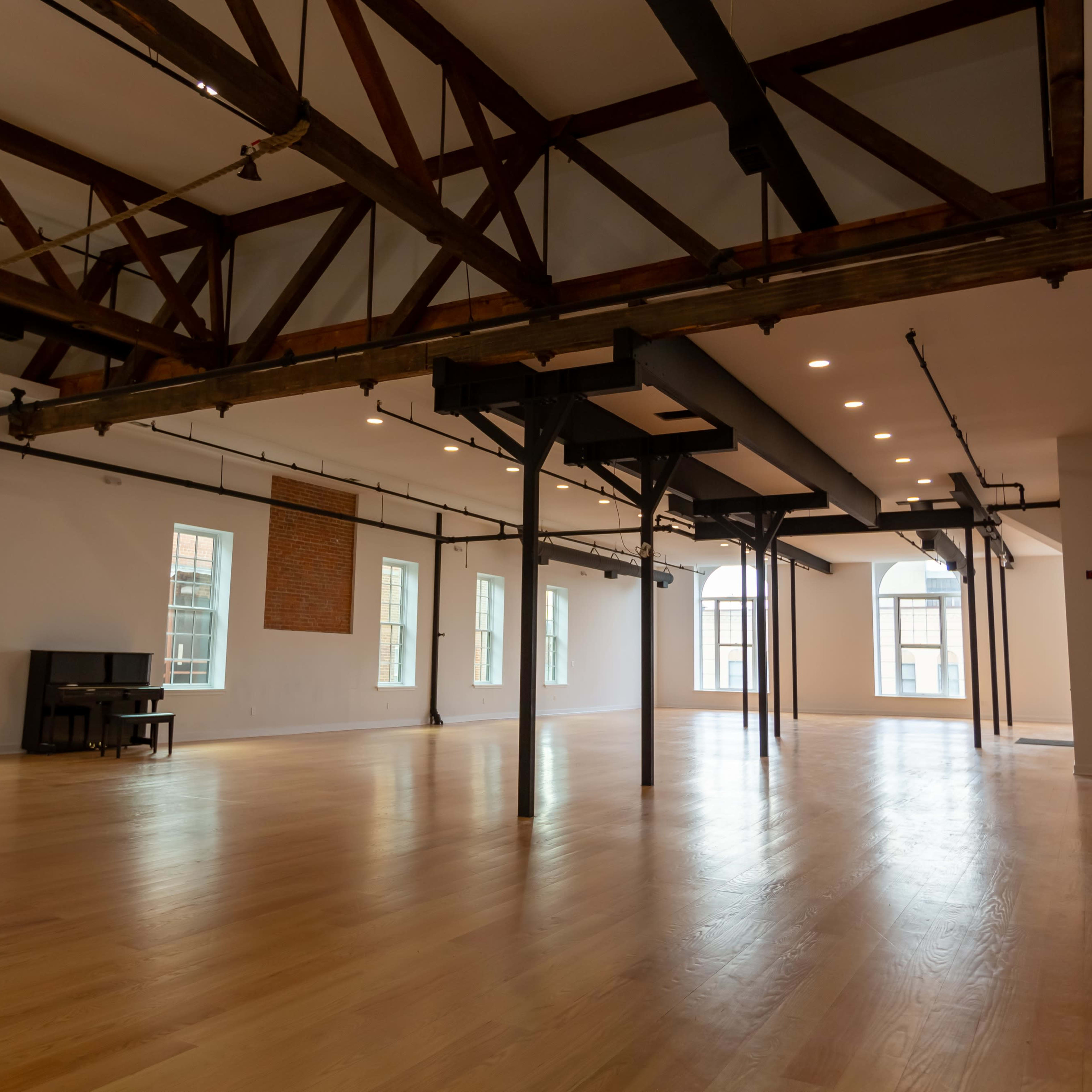 A spacious empty room with wooden flooring, exposed beams, and large windows, featuring a piano in one corner.