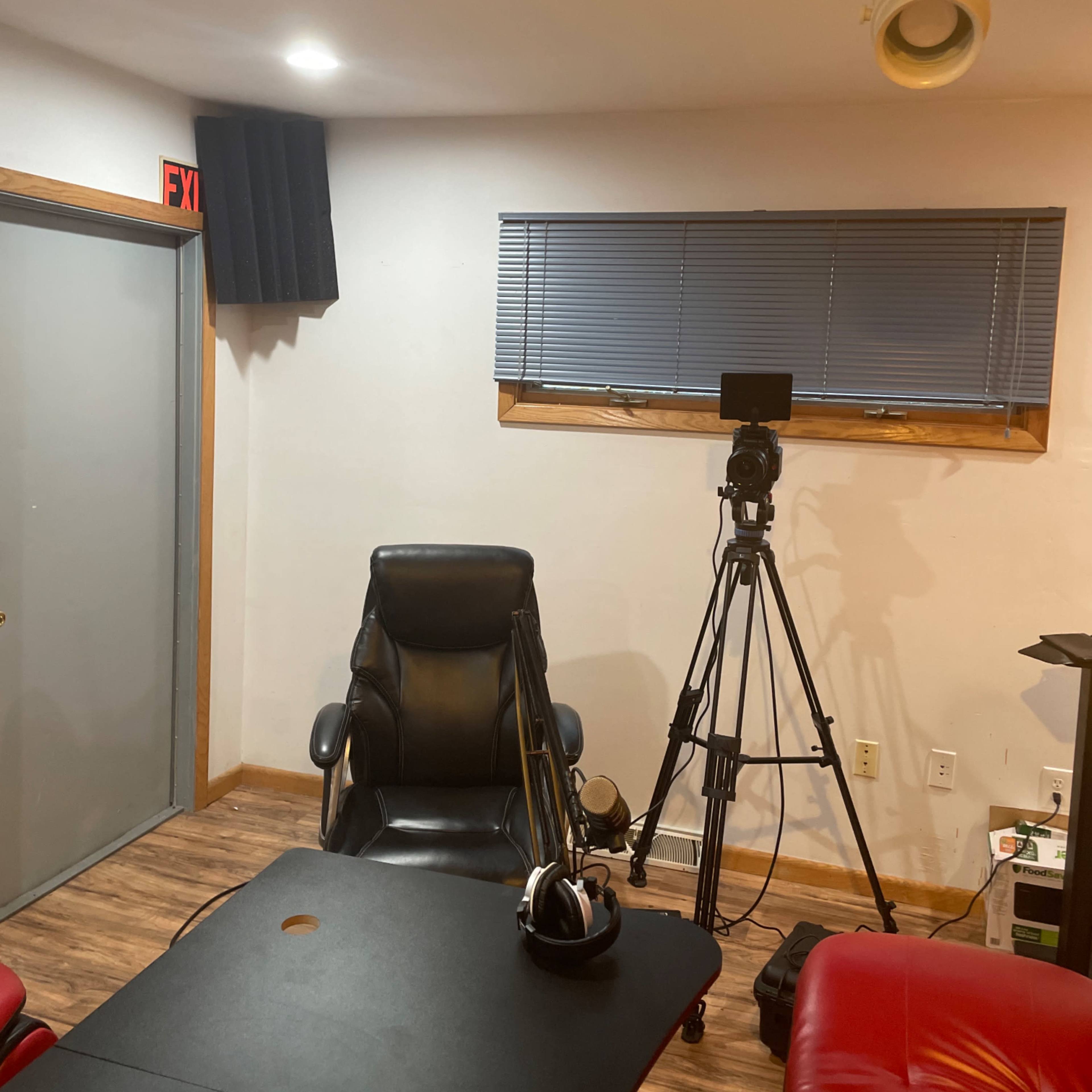 A recording studio with a black chair, a tripod-mounted camera, a desk, and soundproofing panels on the wall.