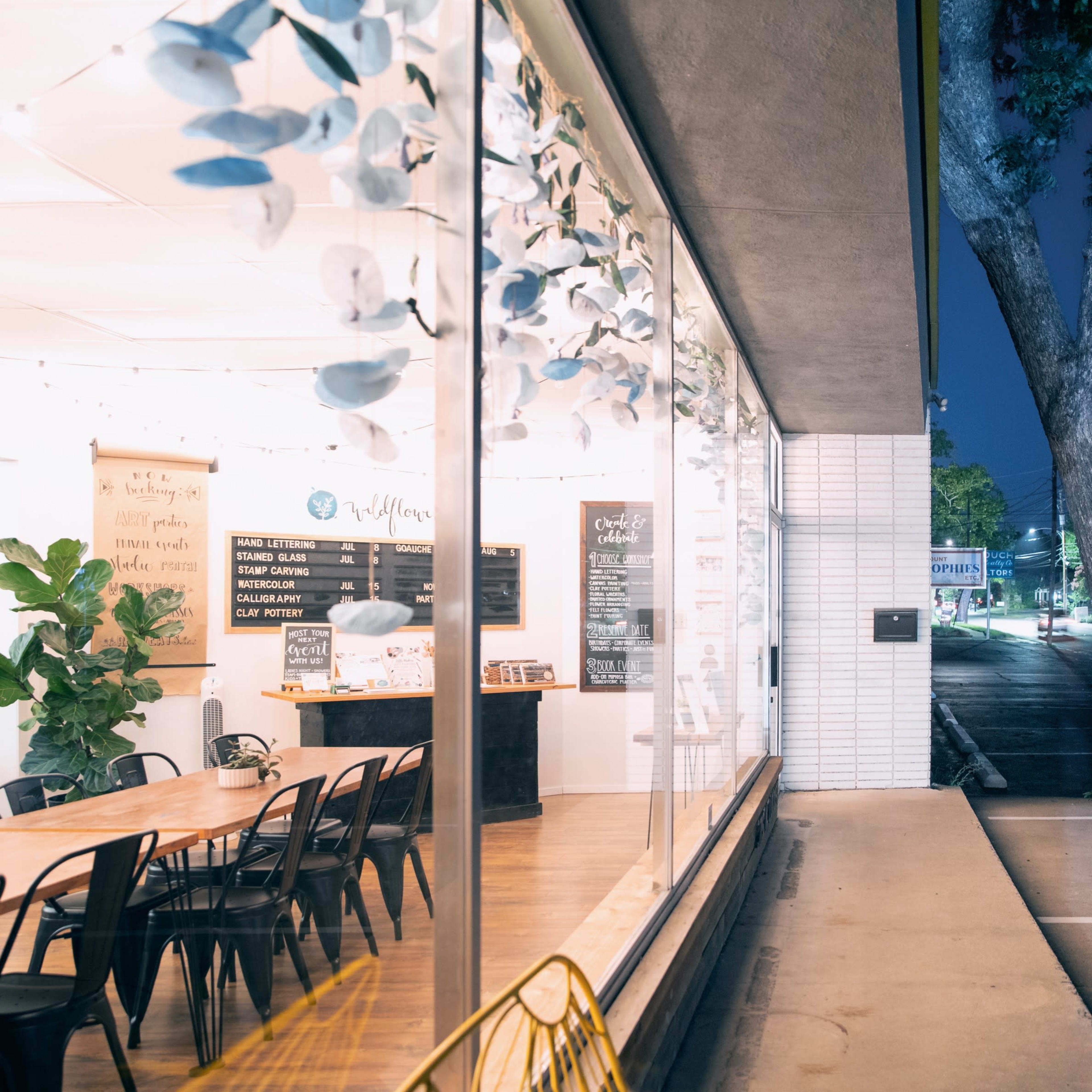 A bright café interior with a large communal table, plants, and a menu board visible through expansive windows at night.
