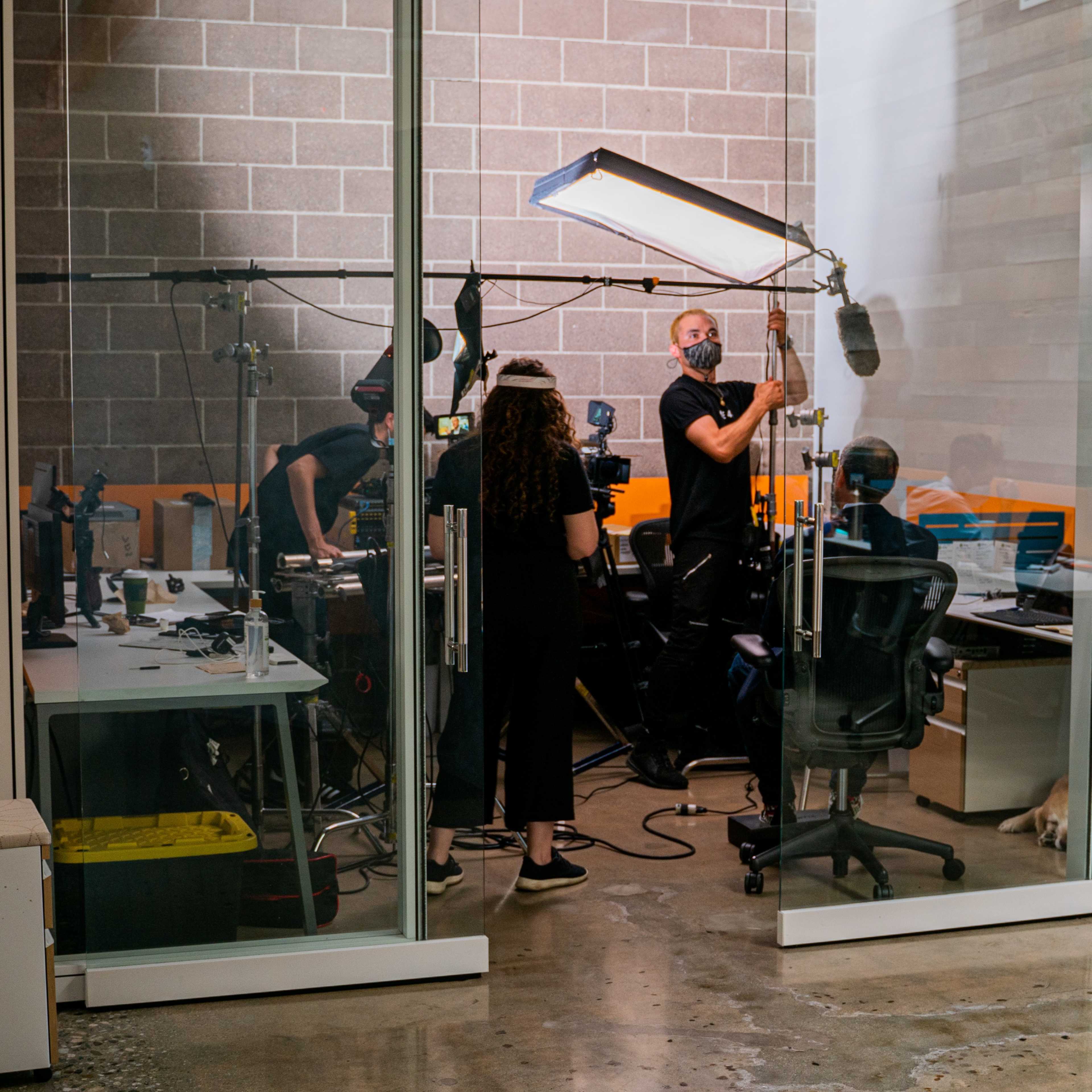 A film crew sets up equipment in a glass-enclosed office space, while a dog rests on the floor nearby.