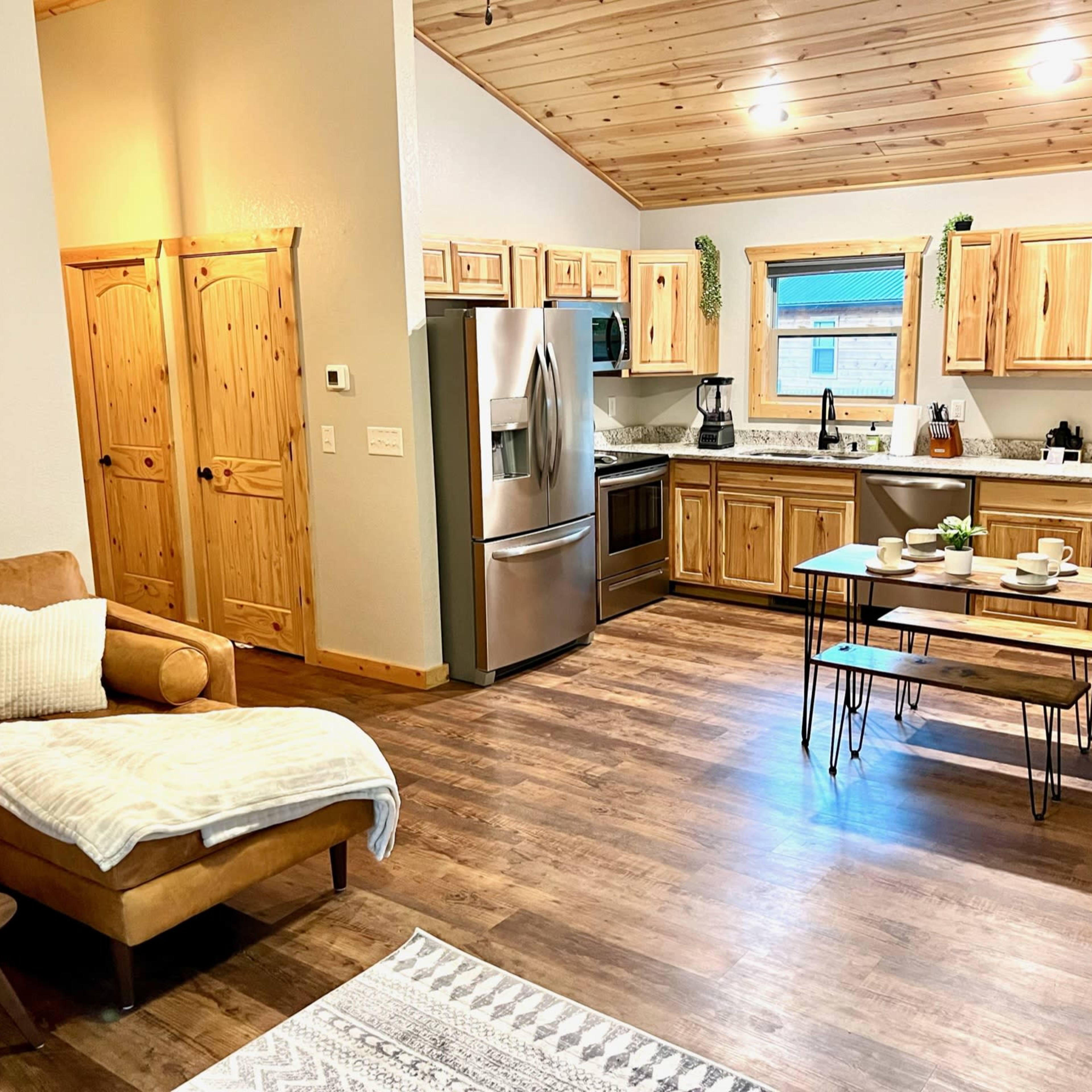 A modern kitchen and living area with wooden cabinetry, stainless steel appliances, a dining table set for four, and a cozy seating area featuring a brown leather couch.