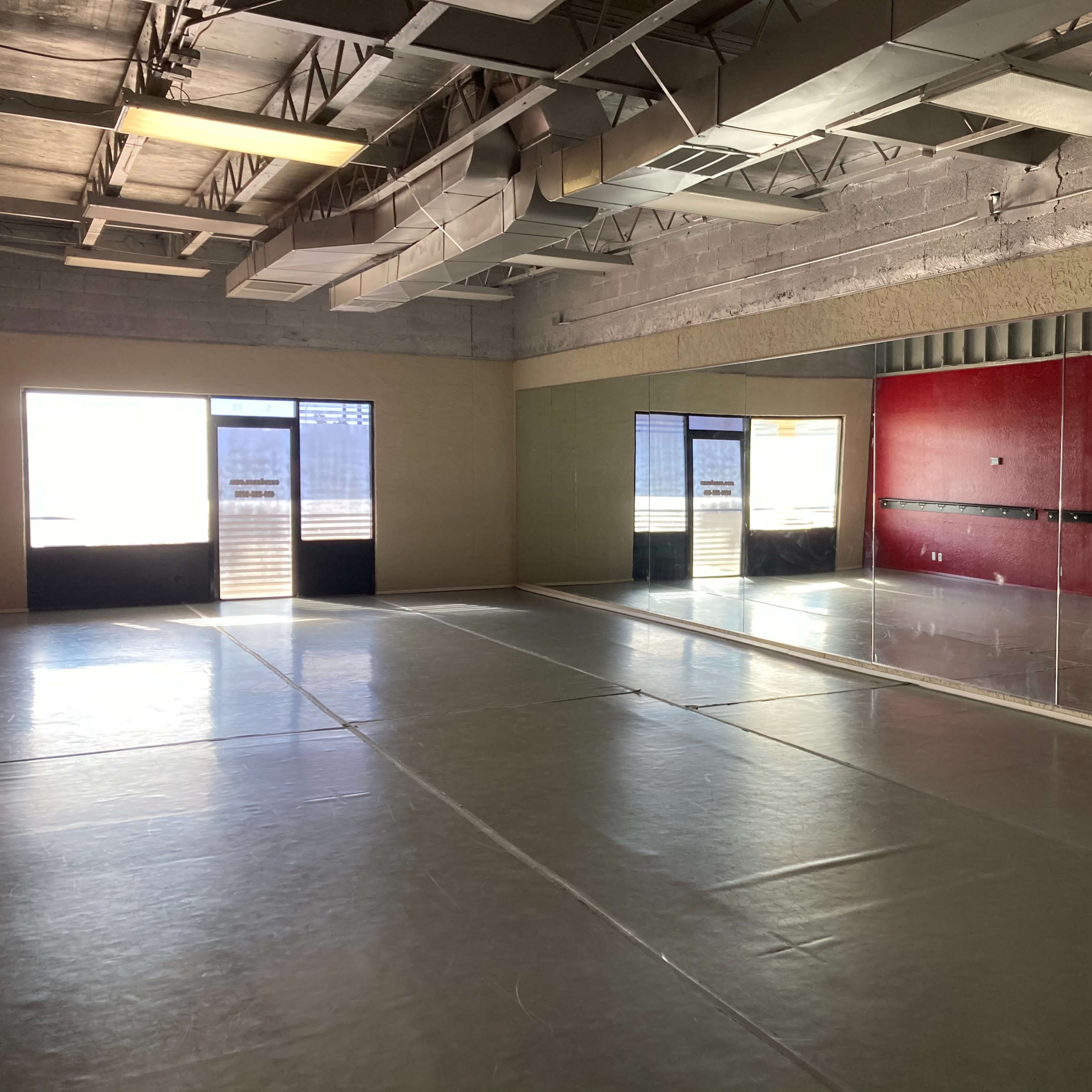 A spacious dance studio with large mirrors, wooden floors, and two sets of glass doors leading to an outside area.